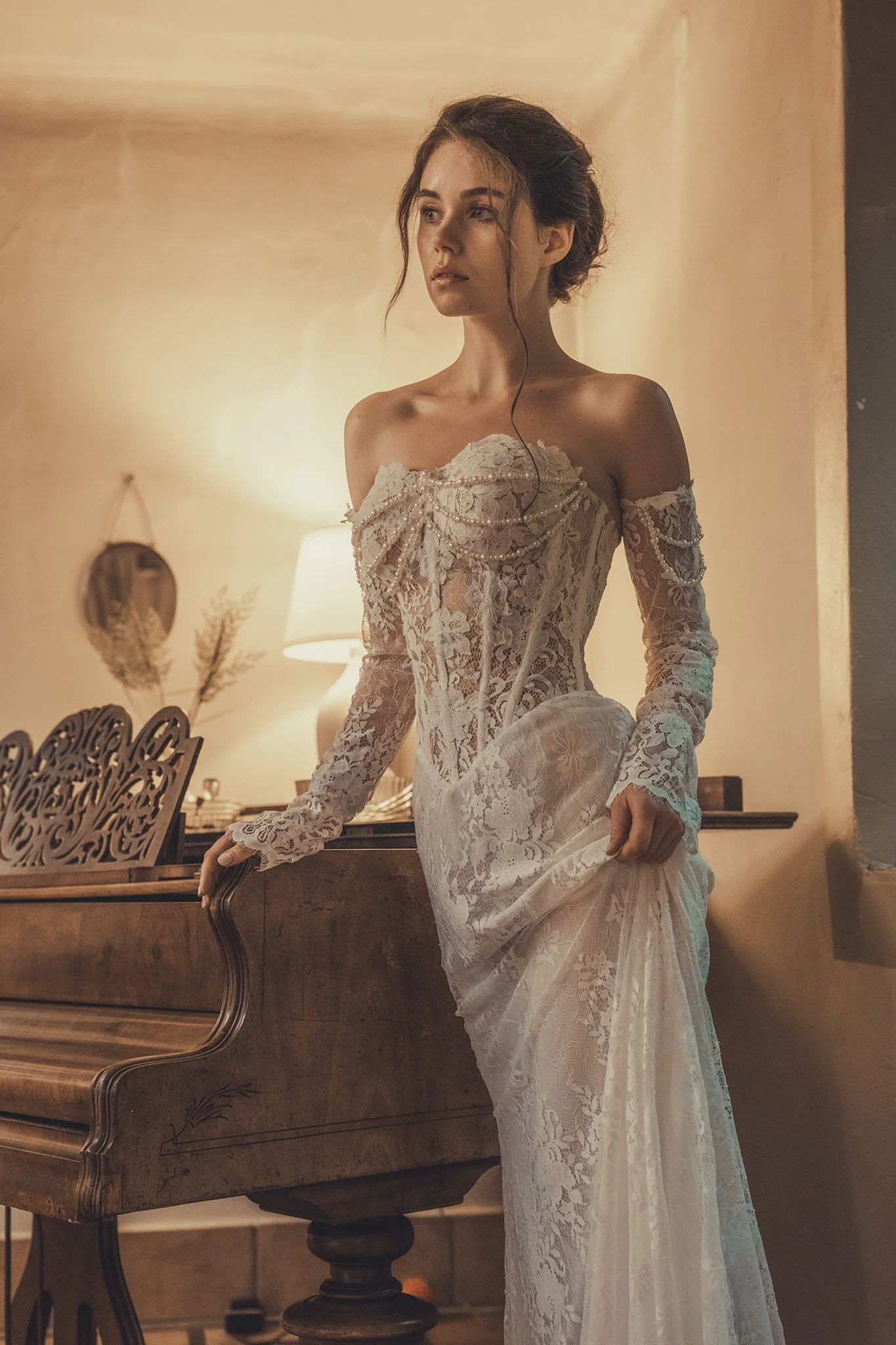 A woman wearing an elegant off-the-shoulder lace wedding dress with long sleeves in a warm-lit room, standing next to a vintage wooden piano.