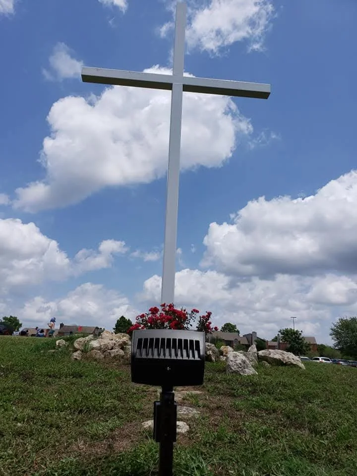 A large white cross mounted on a hill with rocks and flowers at its base, under a partly cloudy blue sky.
