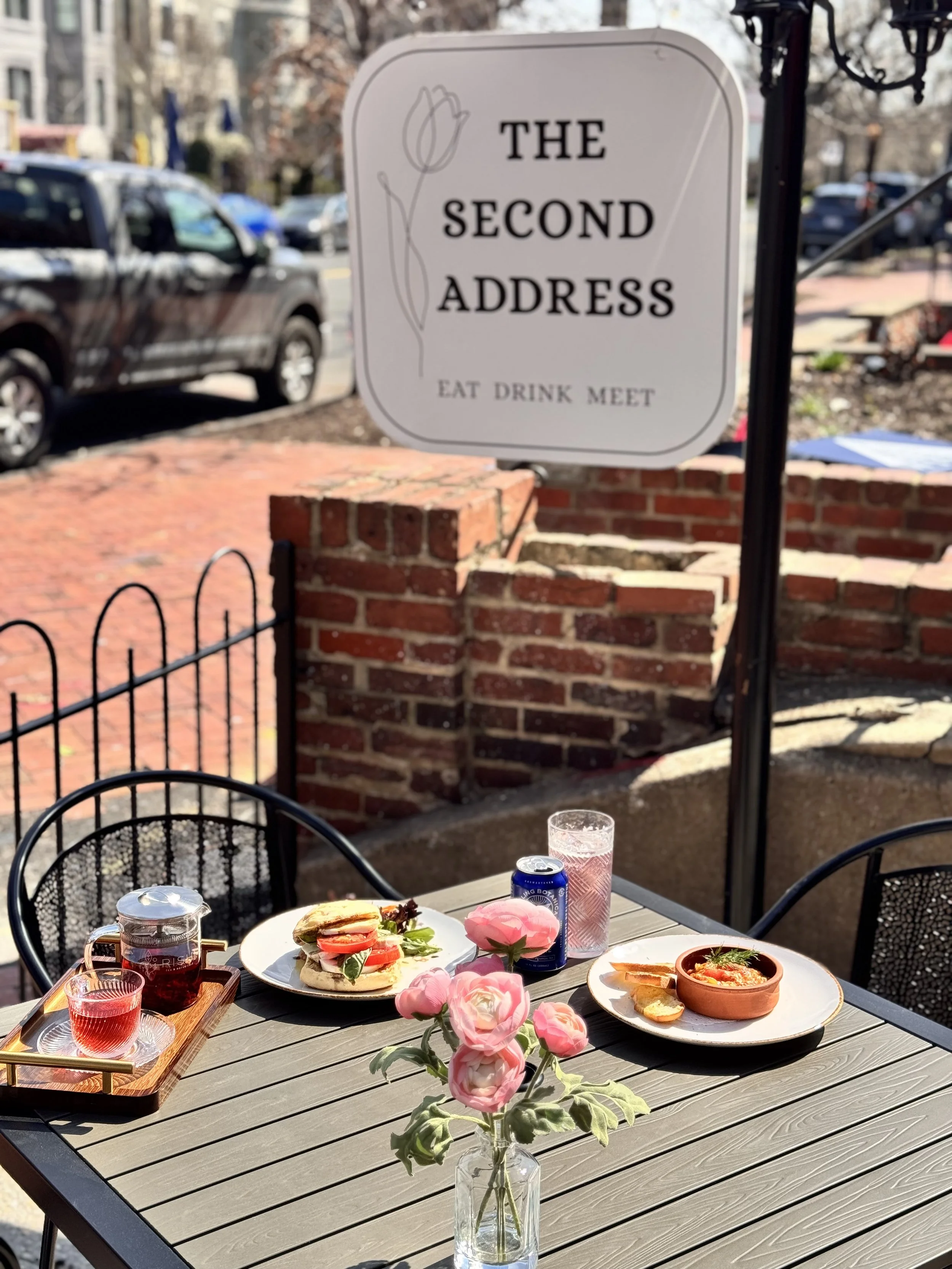 Outdoor cafe table set with a vase of pink flowers, a sandwich, a bowl of soup, a glass of water, a can of soda, and a small glass pitcher of tea. Sign in background reads 'The Second Address, Eat Drink Meet'.