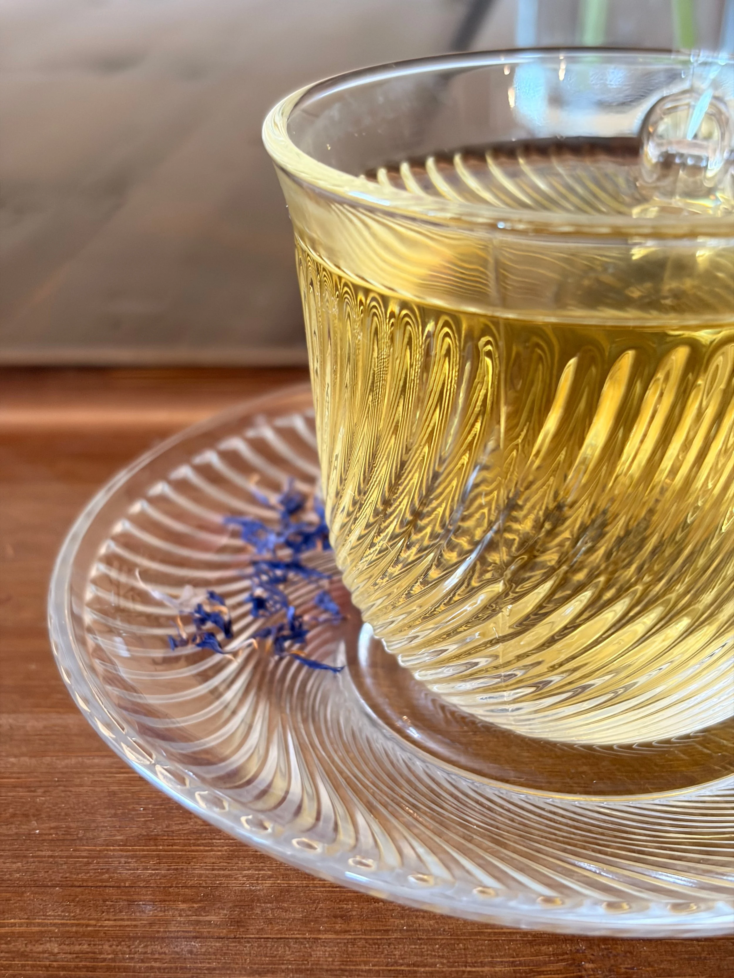 A glass cup filled with a pale yellow liquid, placed on a transparent glass saucer with purple flowers on it, on a wooden surface.