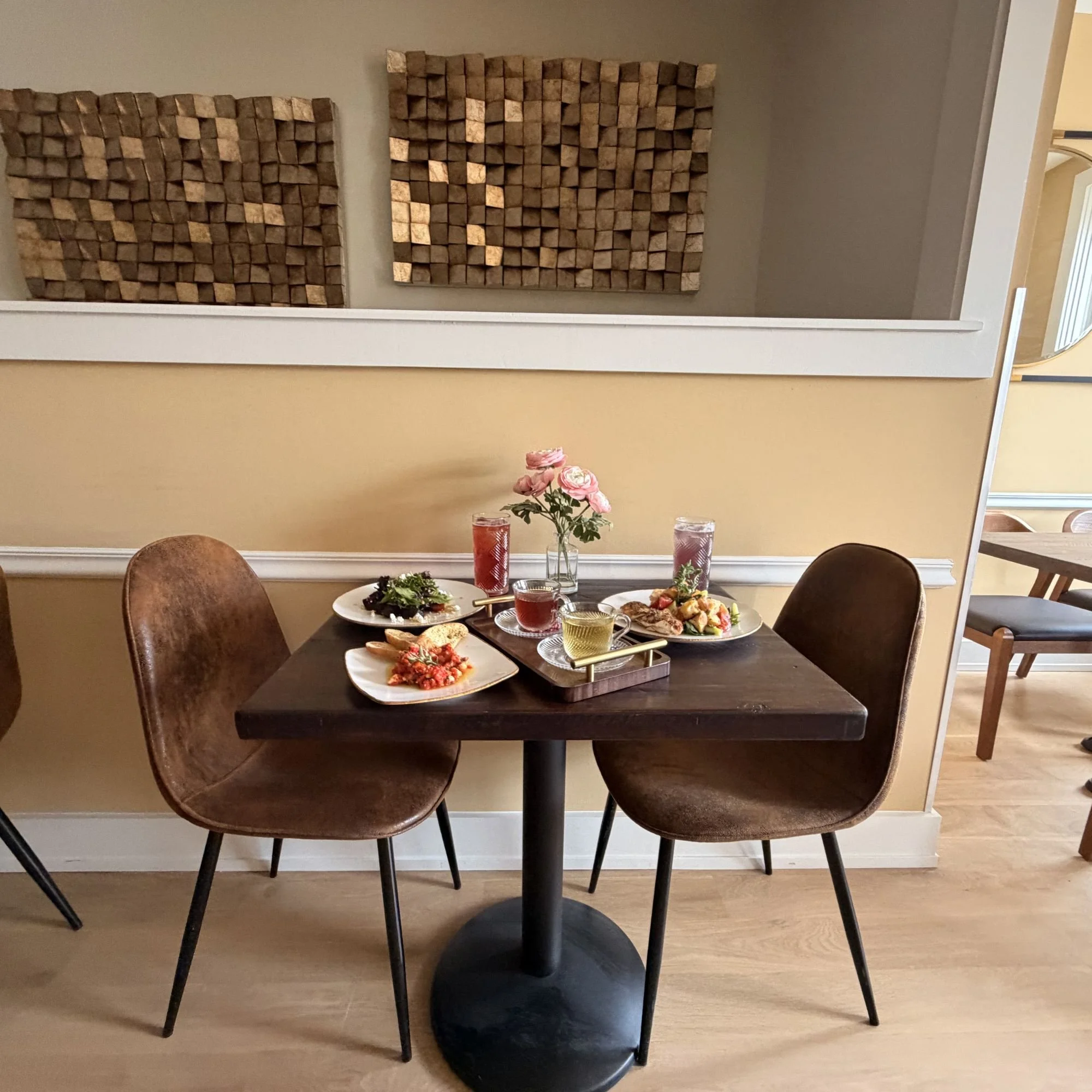 A dining table set for two with various dishes and drinks, a vase of pink roses, and a mirror on a yellow wall, with wooden acoustic panels above.