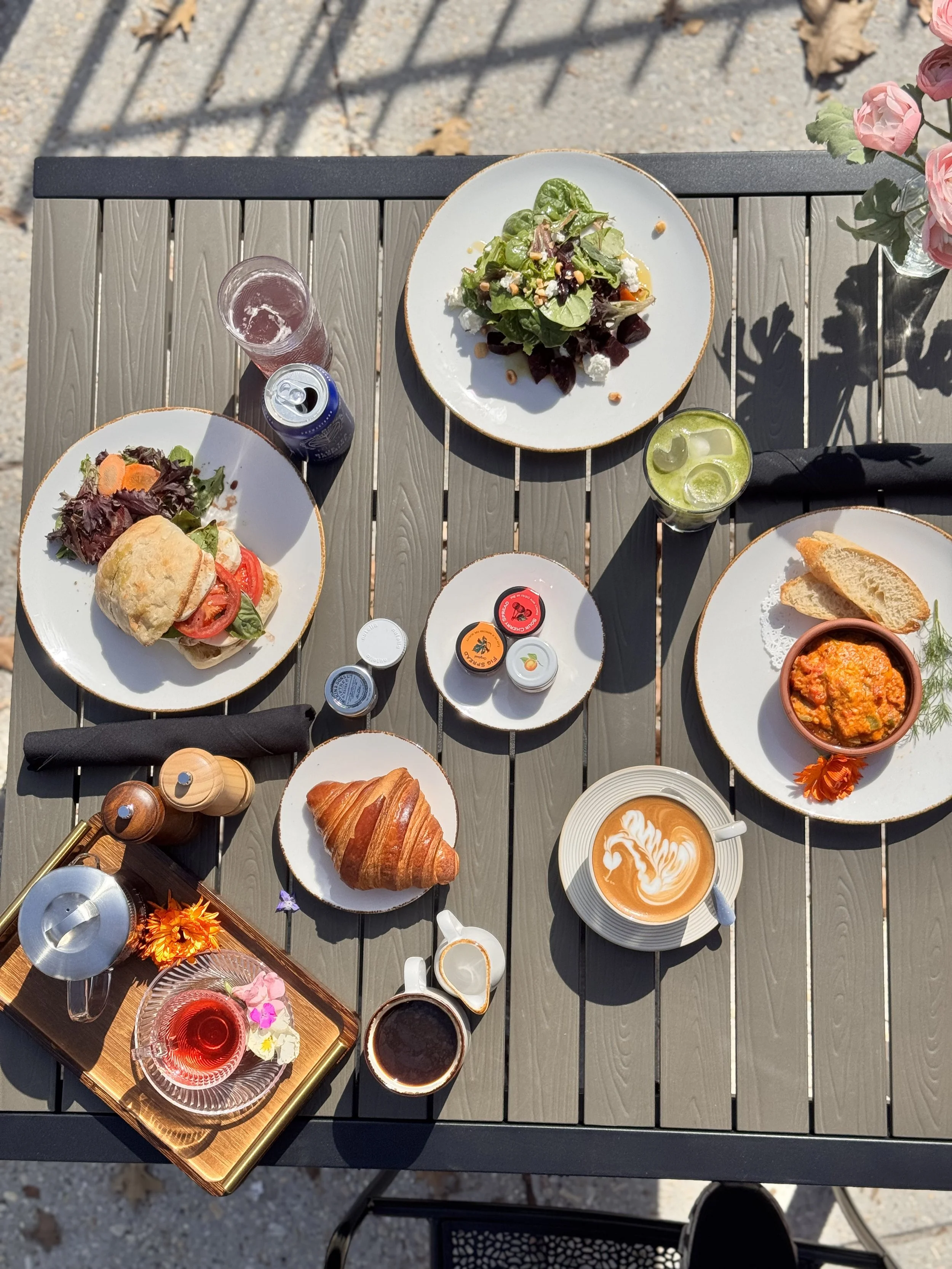 Top-down view of a brunch table with salad, sandwich, croissant, coffee, tea, and drinks, set on a gray outdoor table with flowers on the edge.