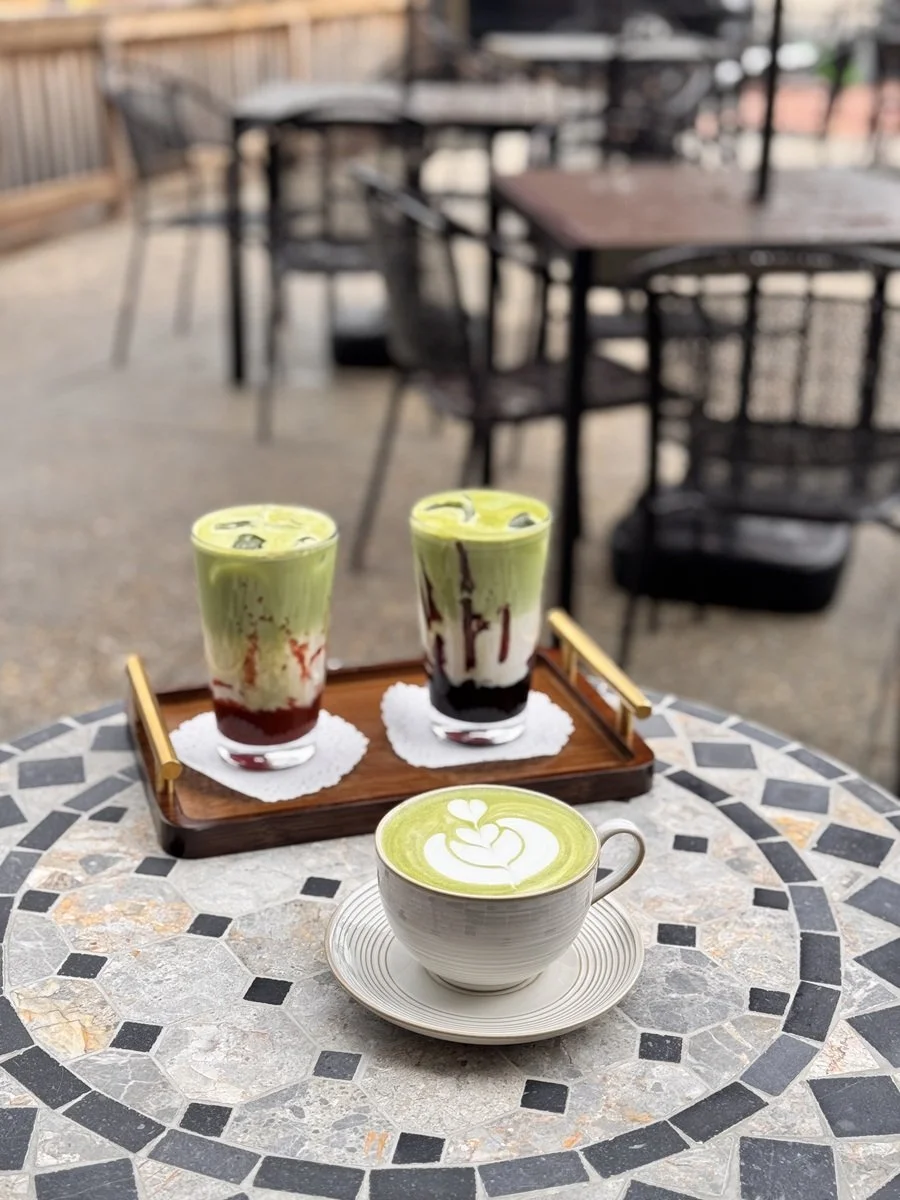 Two layered drinks with green matcha and red syrup, and a matcha latte with latte art on a mosaic table outdoors at a café