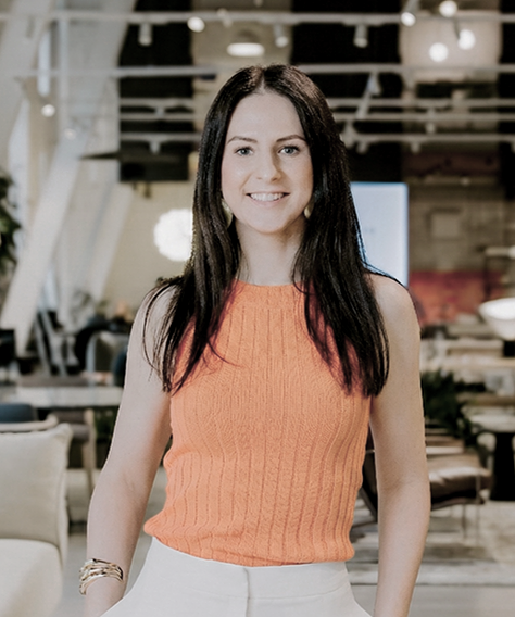 A woman with long dark hair smiling, wearing an orange sleeveless top and white pants, in a modern indoor space with wooden elements and furniture.