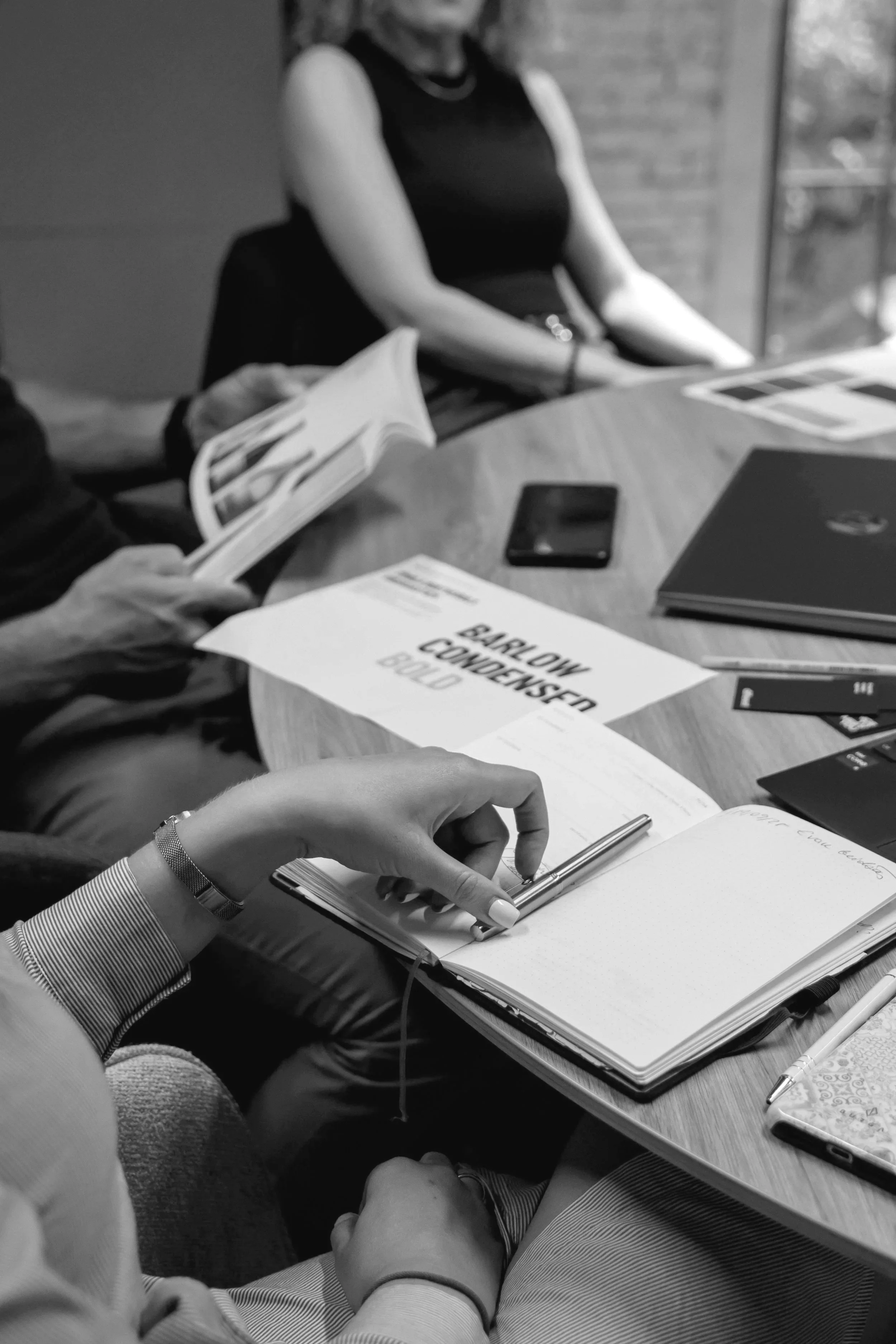 Black and white photo of a meeting with people sitting around a table with notebooks, documents, a smartphone, and pens. One person is writing in a notebook, and another is holding a magazine.
