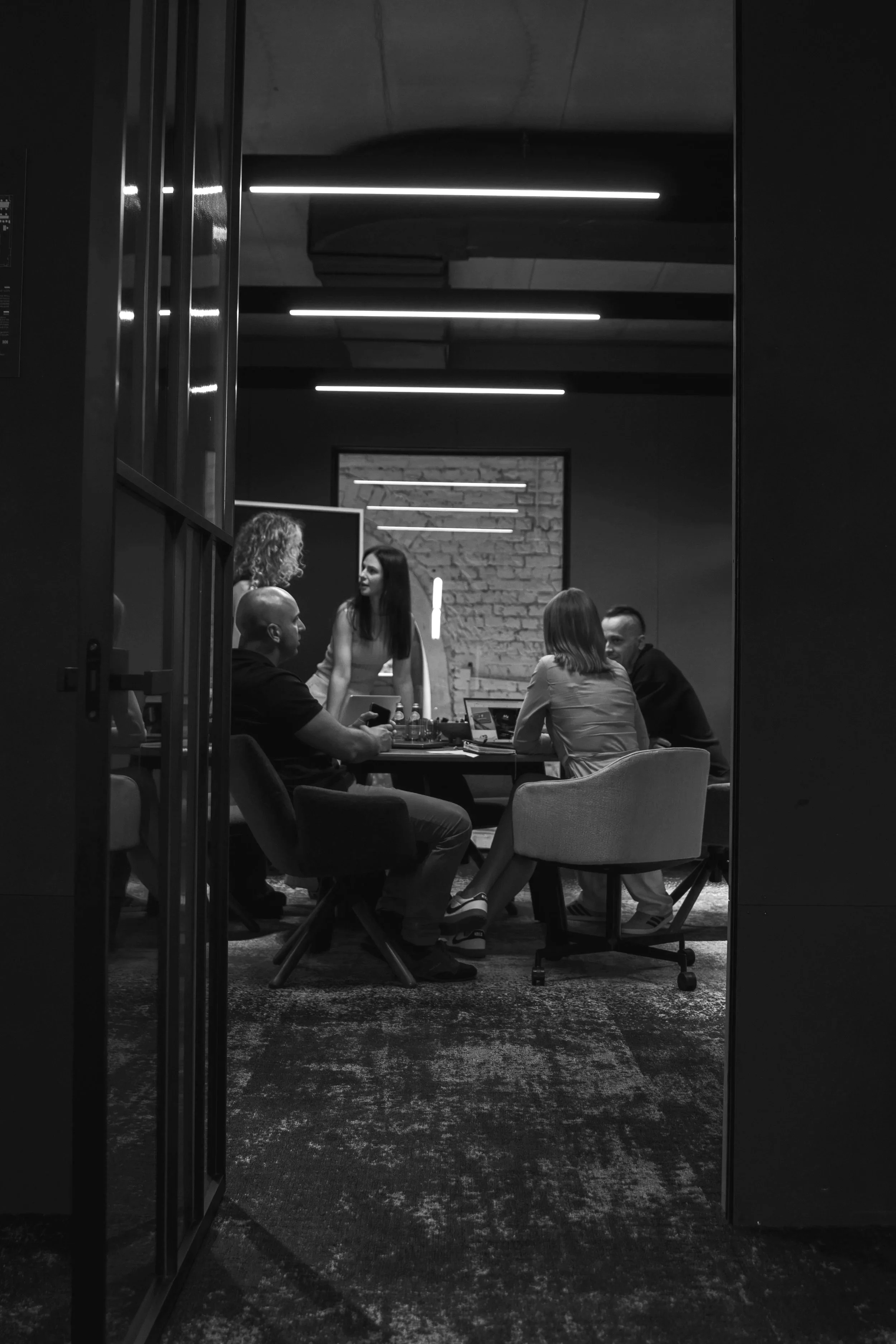 Black and white photo of five people gathered around a table in a modern, industrial-style meeting room. Four are seated and one is standing, engaged in conversation. The room has exposed brick and contemporary lighting fixtures.