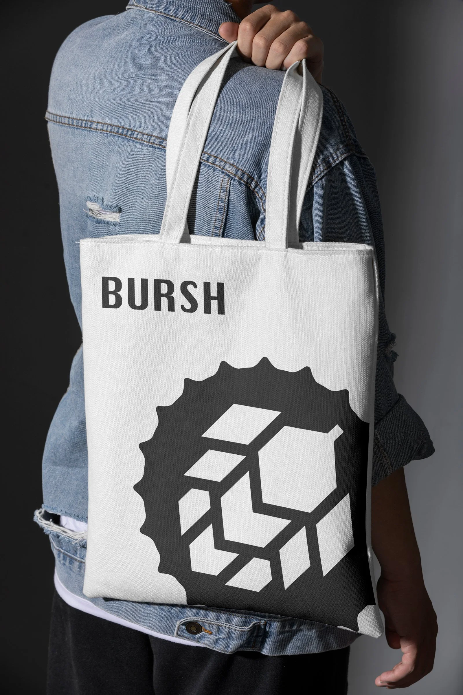 Person wearing a denim jacket carrying a white tote bag with the word 'BURSH' and a geometric cube design on it.