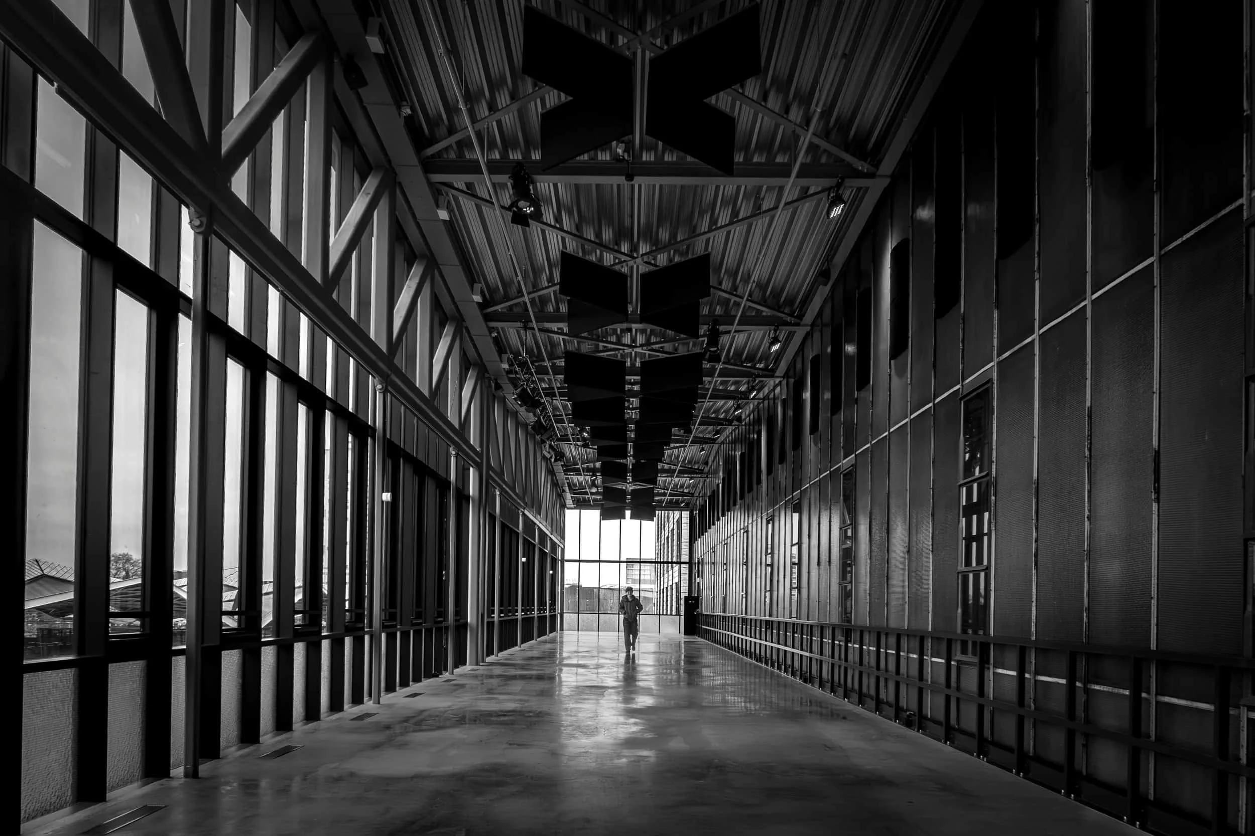 A person walking in a long, modern architectural corridor with glass walls and metal ceiling and panels, black and white photo.