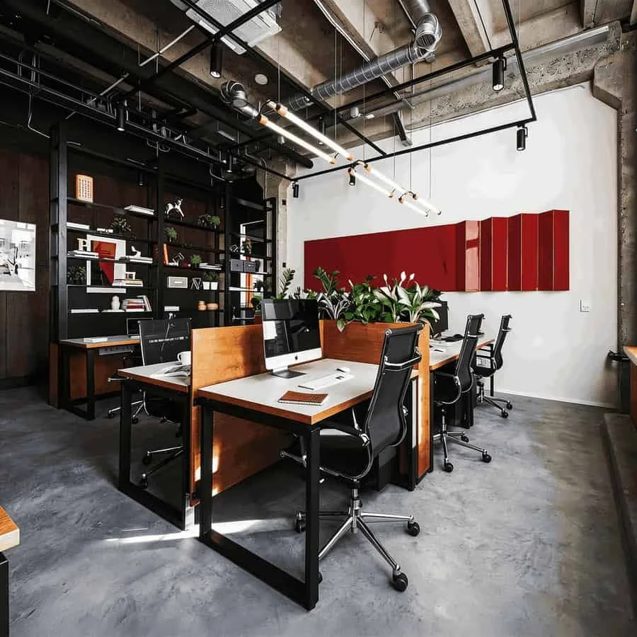 Modern office space with desks, chairs, computers, bookshelves, and green plants, featuring an industrial design with exposed ceiling pipes and concrete walls.