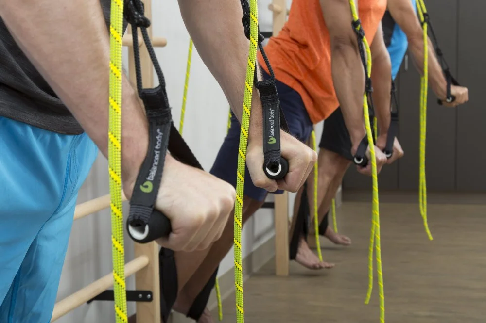 People doing resistance training with ropes, wearing wrist straps labeled 'balanced body' in a fitness studio.