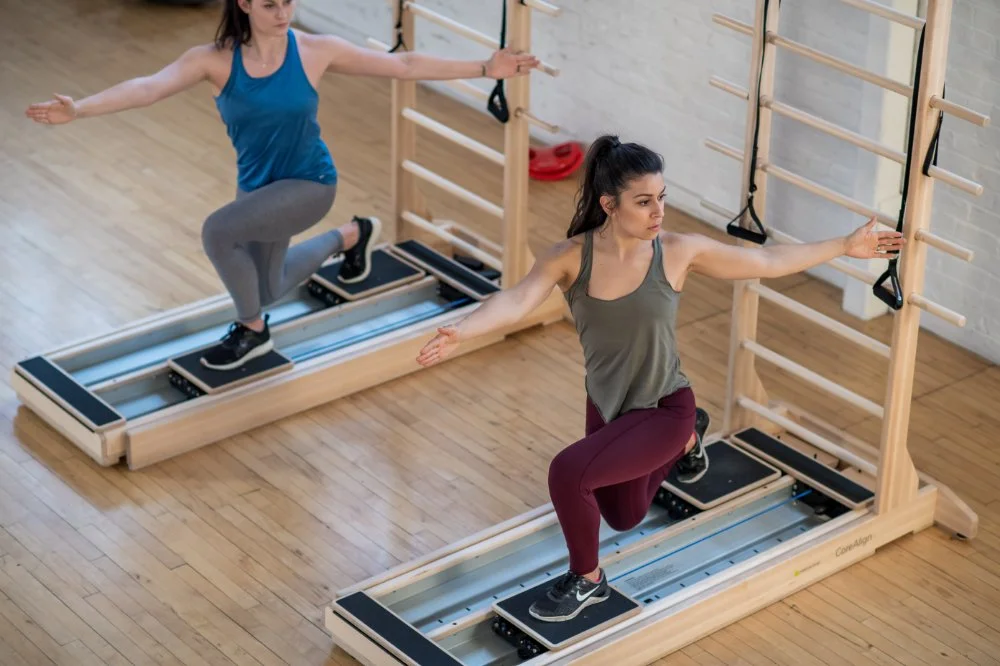 Two women exercising on reformer Pilates machines in a fitness studio, with one woman in a green tank top and maroon leggings kneeling and extending her arms, and the other woman in a blue tank top and gray leggings standing behind her, both engaging in a workout session.