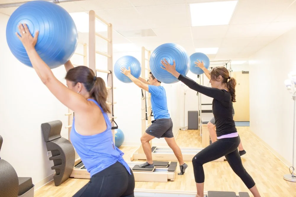 Group of people participating in a fitness class using large blue exercise balls and platforms in a gym.