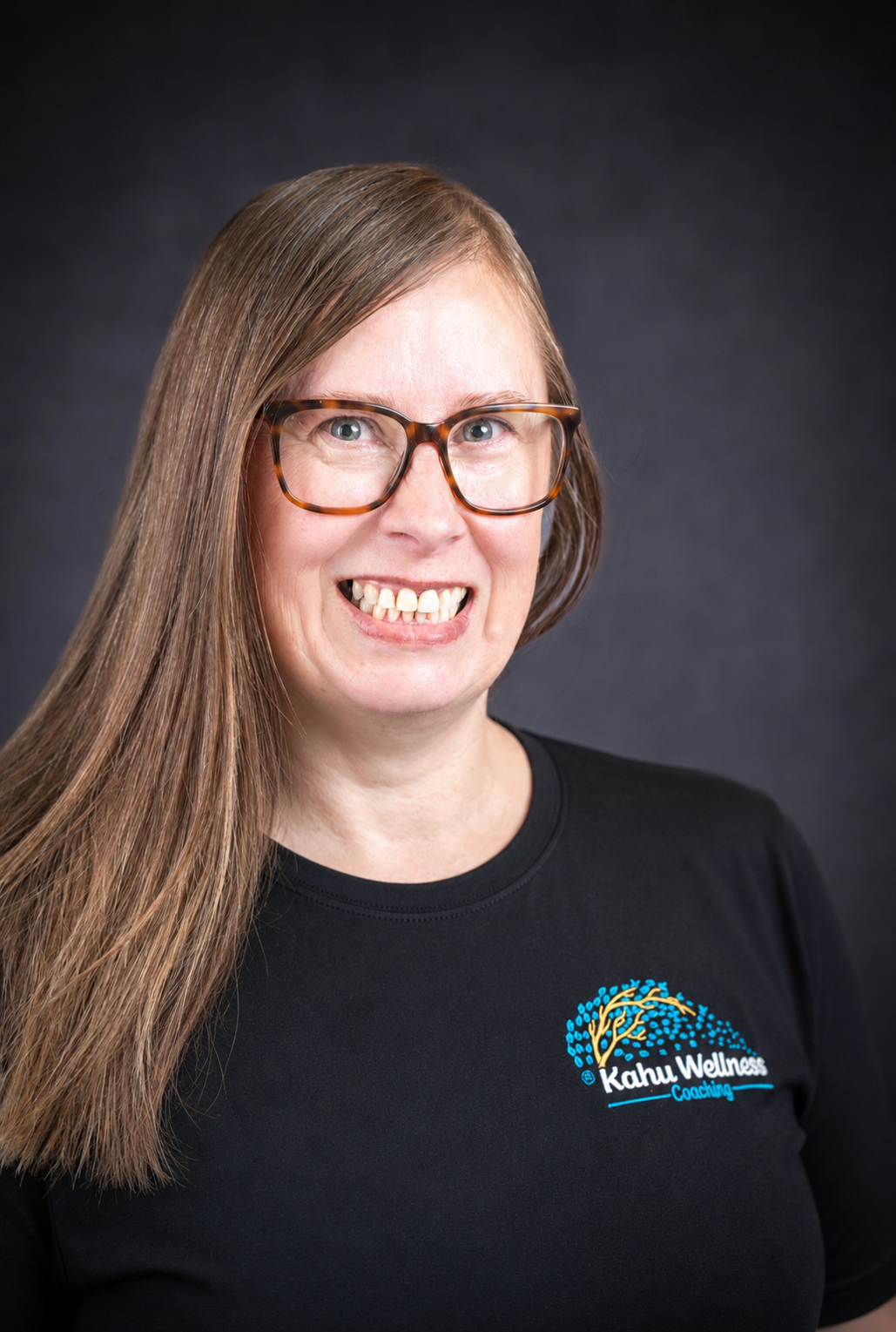 A woman with long brown hair, glasses, and a black shirt with a logo that reads 'Kahu Wellness Coaching,' smiling against a dark background.
