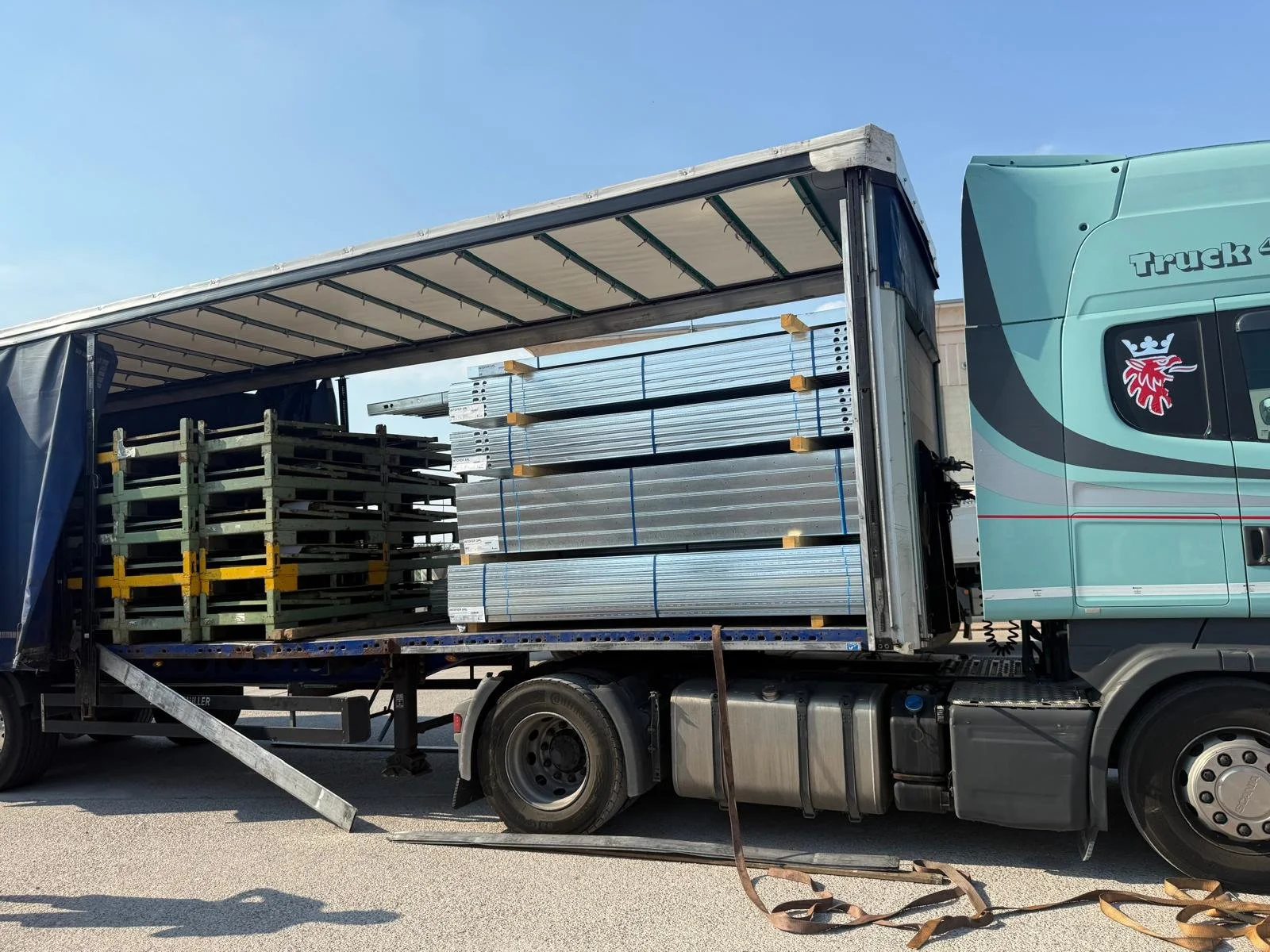 Un camion trasporta impalcature di metallo e scaffali di legno, parcheggiato all'aperto sotto un cielo azzurro.