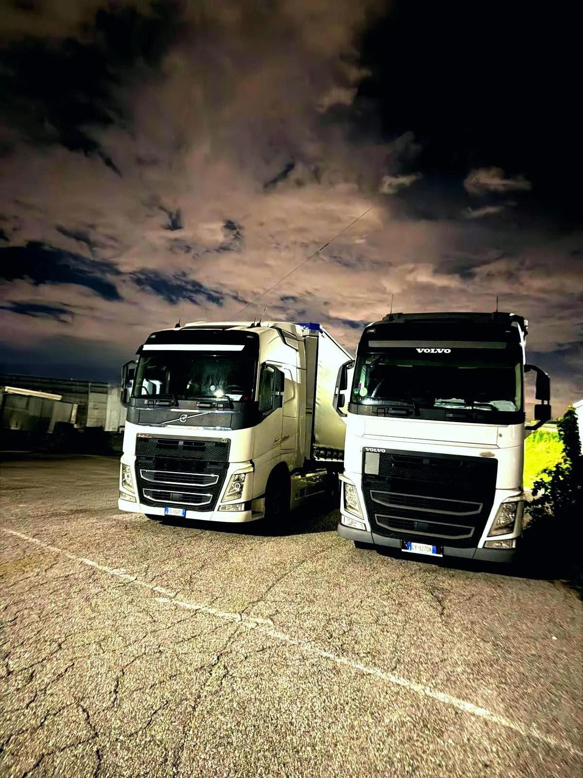 Due camion Volvo parcheggiati su una strada all'aperto di notte, con cielo nuvoloso sopra.