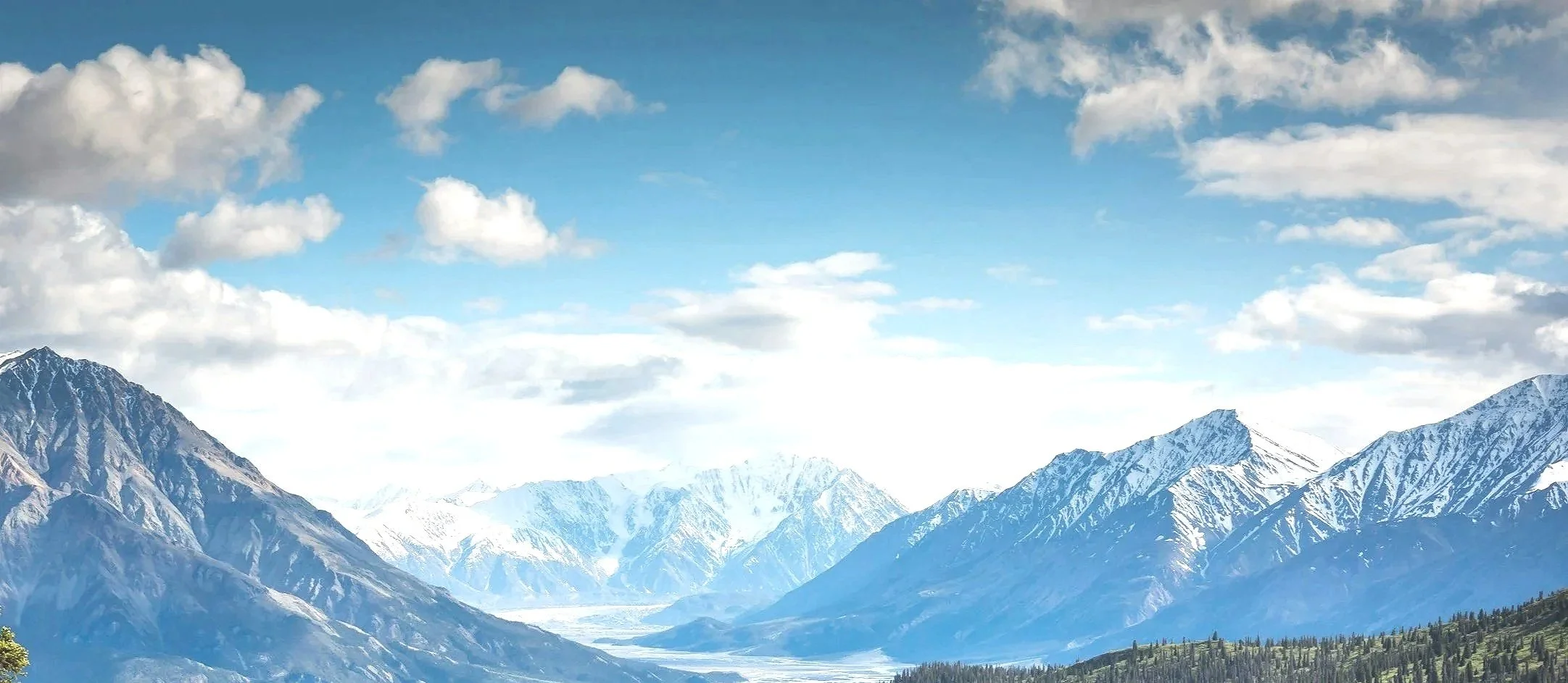 Snow-capped mountains under a partly cloudy blue sky with some green trees at the base.