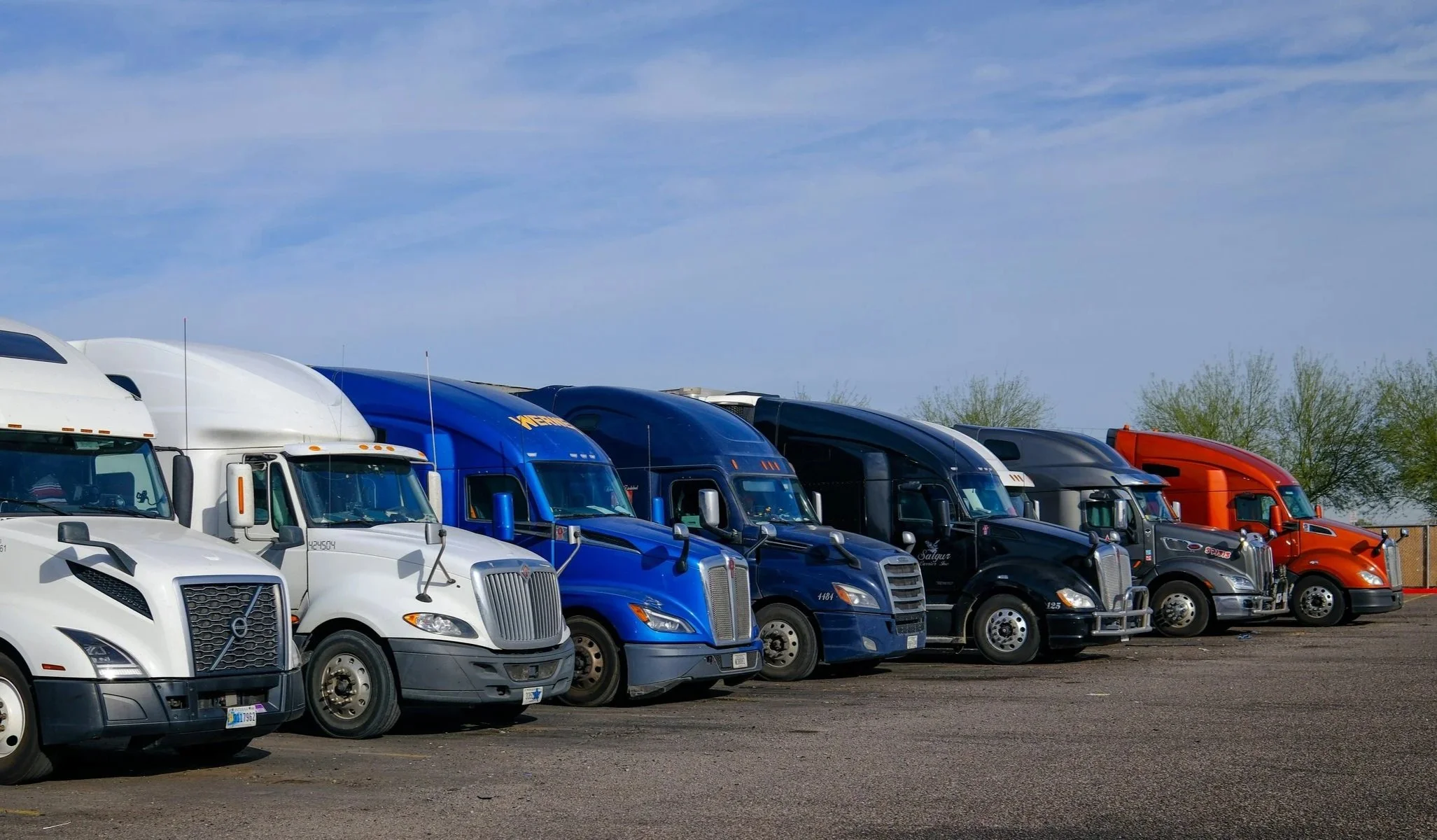 Row of various colored semi-trucks parked on a lot under a partly cloudy sky.