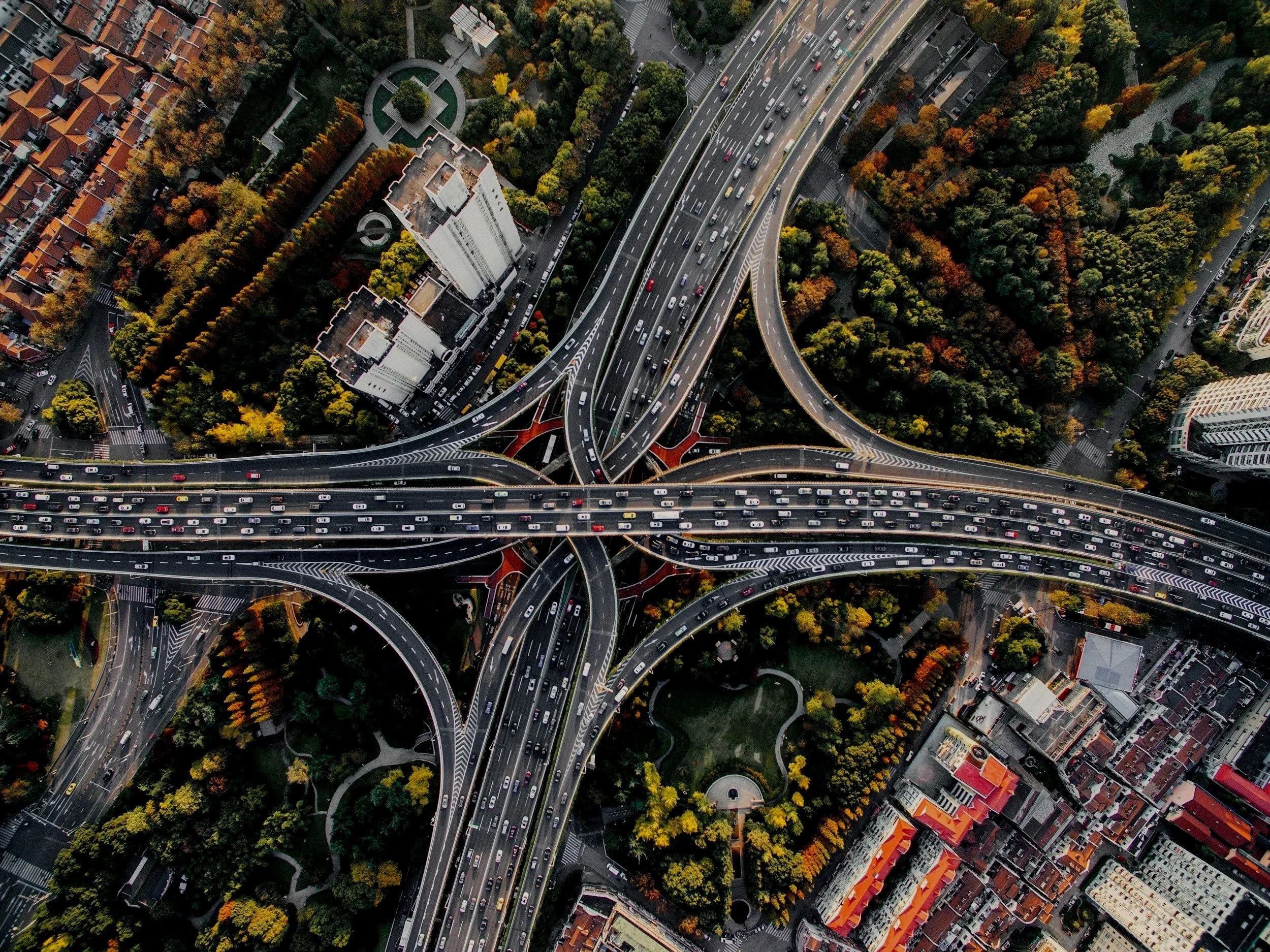 An aerial view of a multi-level highway interchange surrounded by city buildings, residential areas, and trees with autumn foliage.