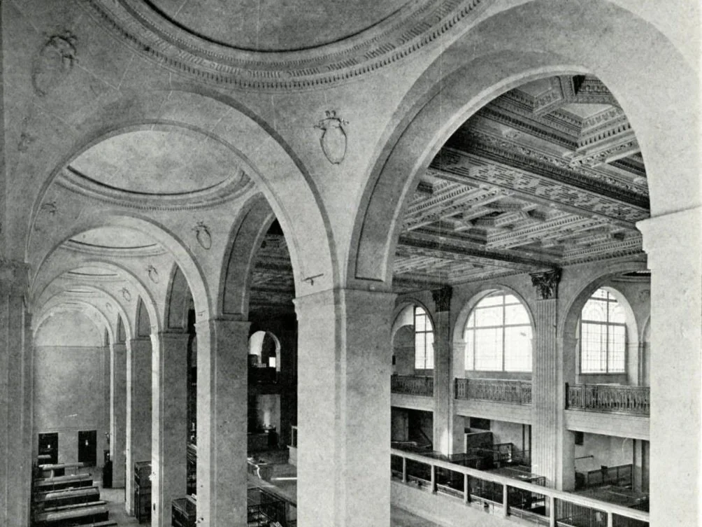 Black and white photo of an ornate interior space with large arched windows, decorative ceiling, and balconies overlooking the lower level.