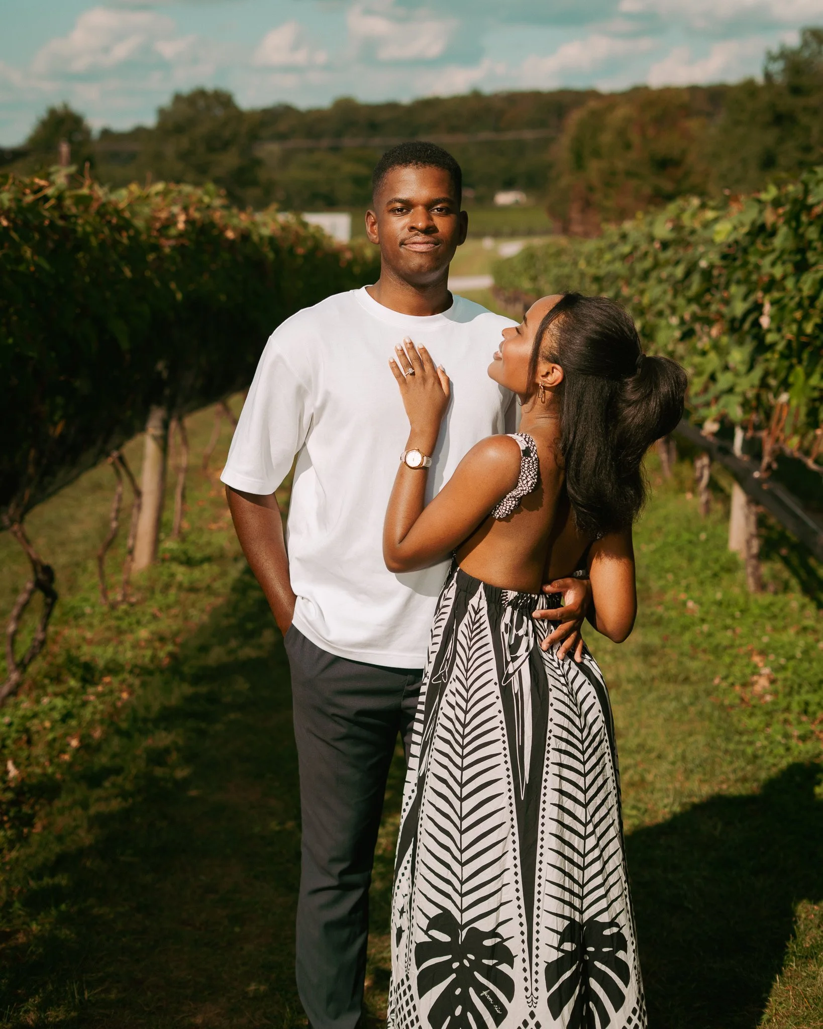 A couple standing close together in a vineyard with lush green vines in the background. The woman is looking up at the man, touching his chest with her hand, wearing a patterned dress. The man is looking straight ahead, dressed in a white t-shirt and dark pants.