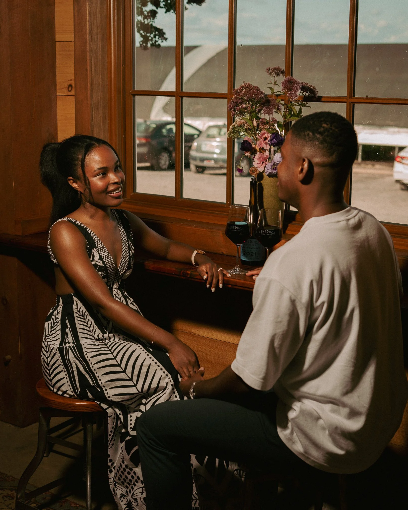 A woman and a man sitting and talking at a wooden bar near a window with two glasses of red wine and a vase with purple flowers.