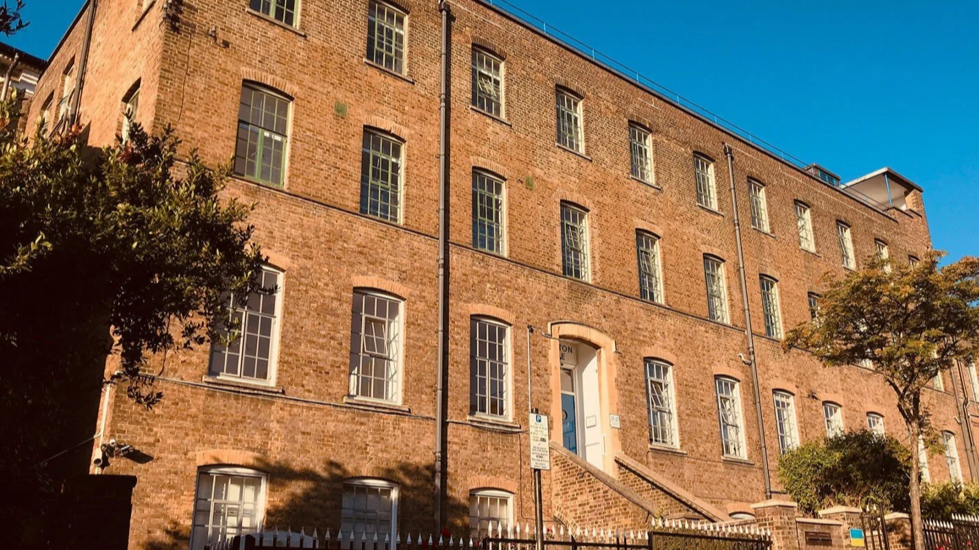 A brick building with multiple windows, a staircase leading to a blue door, and two trees in front under a clear blue sky.