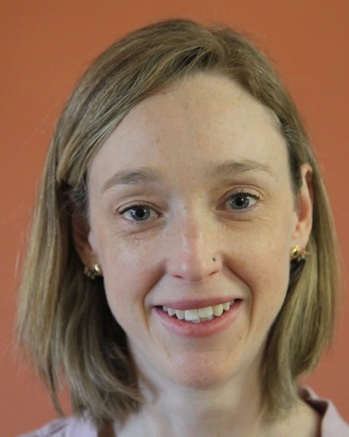 Close-up of a young woman with shoulder-length blonde hair, blue eyes, wearing small earrings and a nose piercing, smiling at the camera against a warm, orange background.