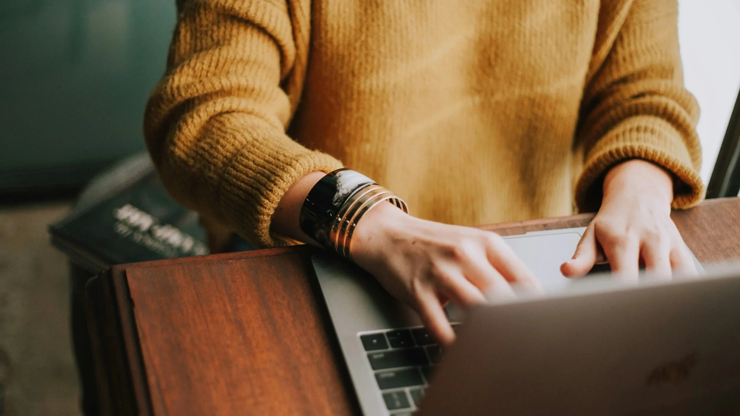 Person wearing a mustard-colored sweater and a black-and-gold bracelet typing on a MacBook laptop at a wooden desk.