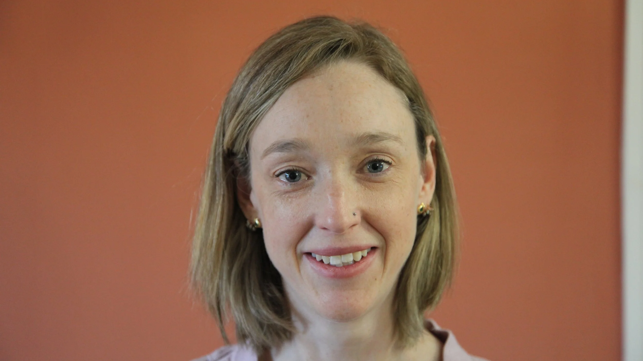 Close-up of a woman with light brown hair, blue eyes, wearing gold earrings, smiling, in front of a reddish-orange background.