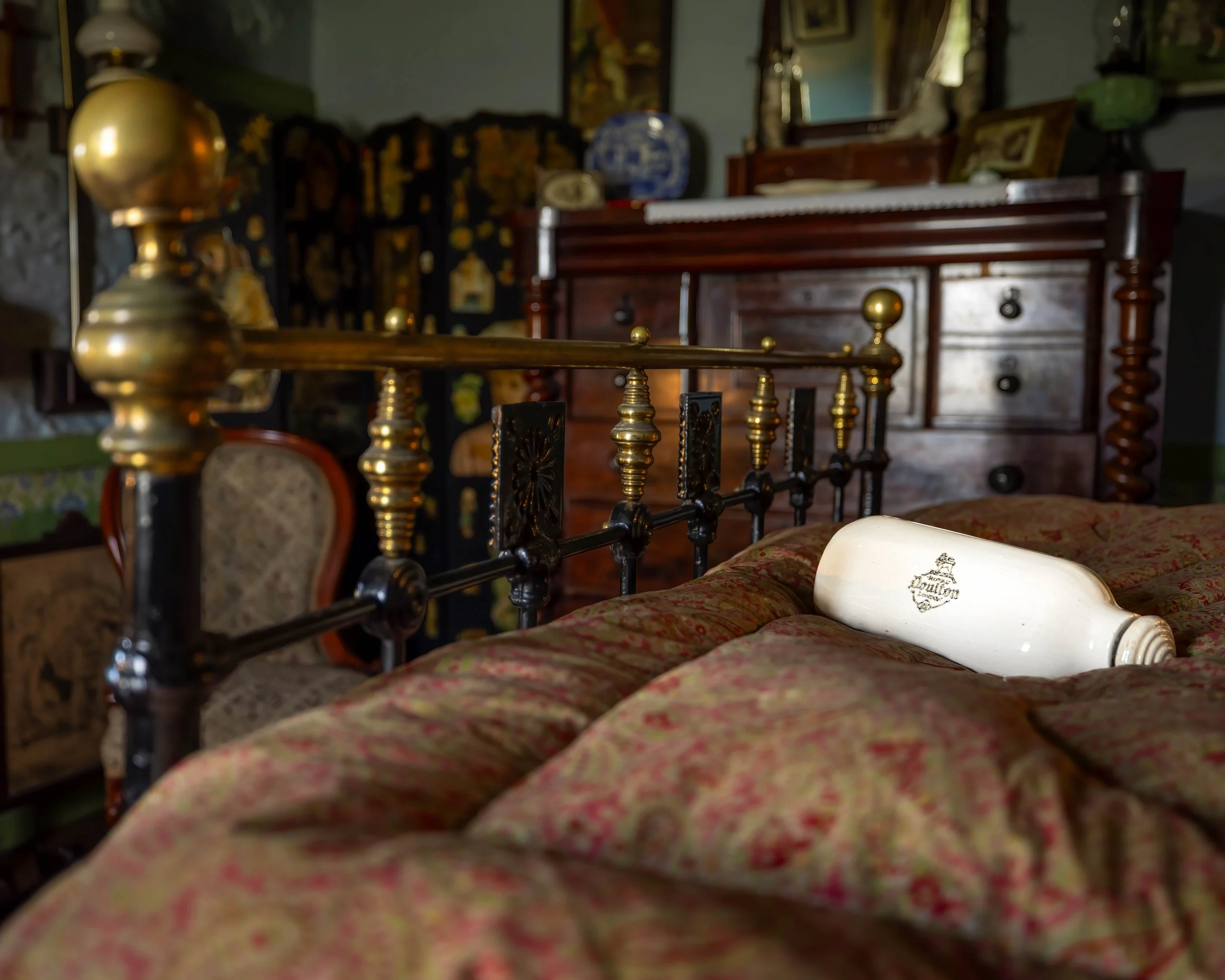 Boloquoy Victorian Farm & Watermill. An antique bed with a patterned bedspread, a white cylinder-shaped object branded 'LONDON' resting on it, and a dark wooden dresser in the background, inside a vintage-style room with various decorative items.