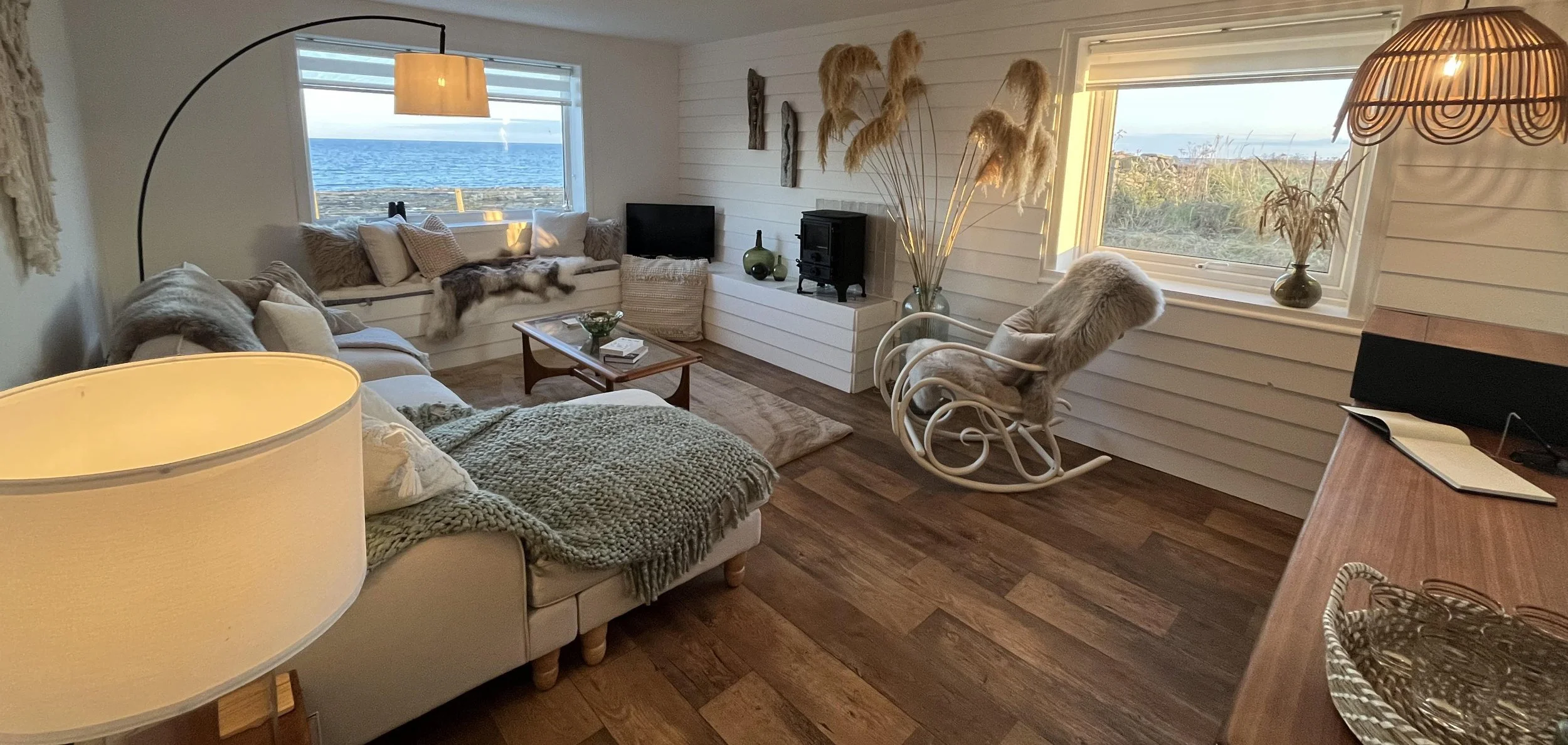 Coastal living room with large windows showing ocean view, cream-colored furniture with cozy throws and pillows, a rocking chair with faux fur, decorative dried plants, and soft ambient lighting.