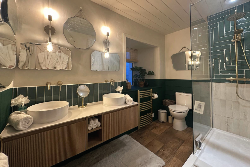 Modern bathroom with double vessel sinks, round mirrors, wall-mounted lights, a toilet, a walk-in shower, greenery on the windowsill, wooden cabinetry, and decorative tiling.