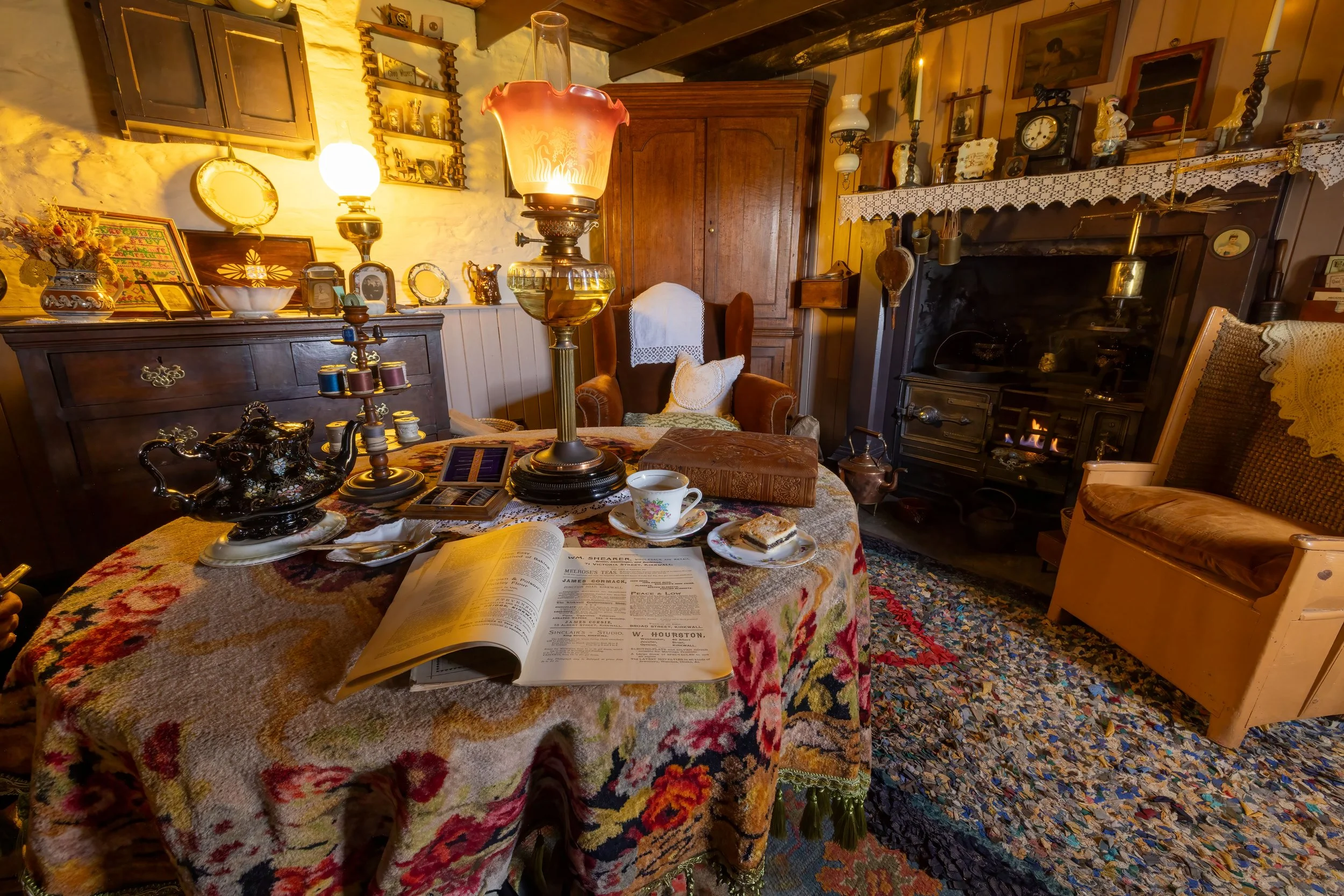 A cozy vintage living room with a round table covered with a floral tablecloth, holding a teapot, a cup and saucer, an open book, and snacks. The room has antique furniture, lamps, a fireplace with a lit fire, and various decorative items and pictures on the walls.