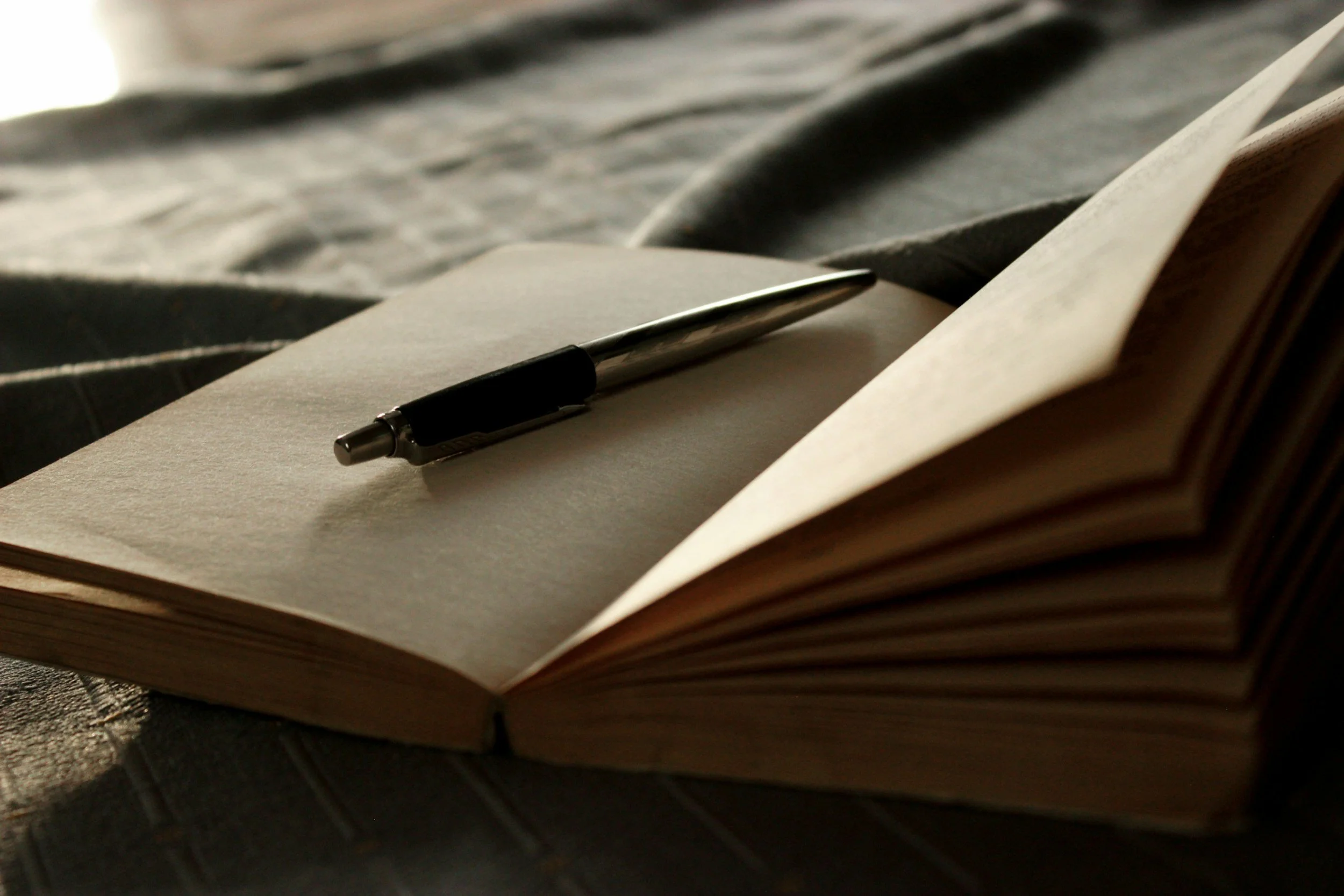 Open notebook with blank pages and a black pen resting on it, on a textured surface with fabric in the background.