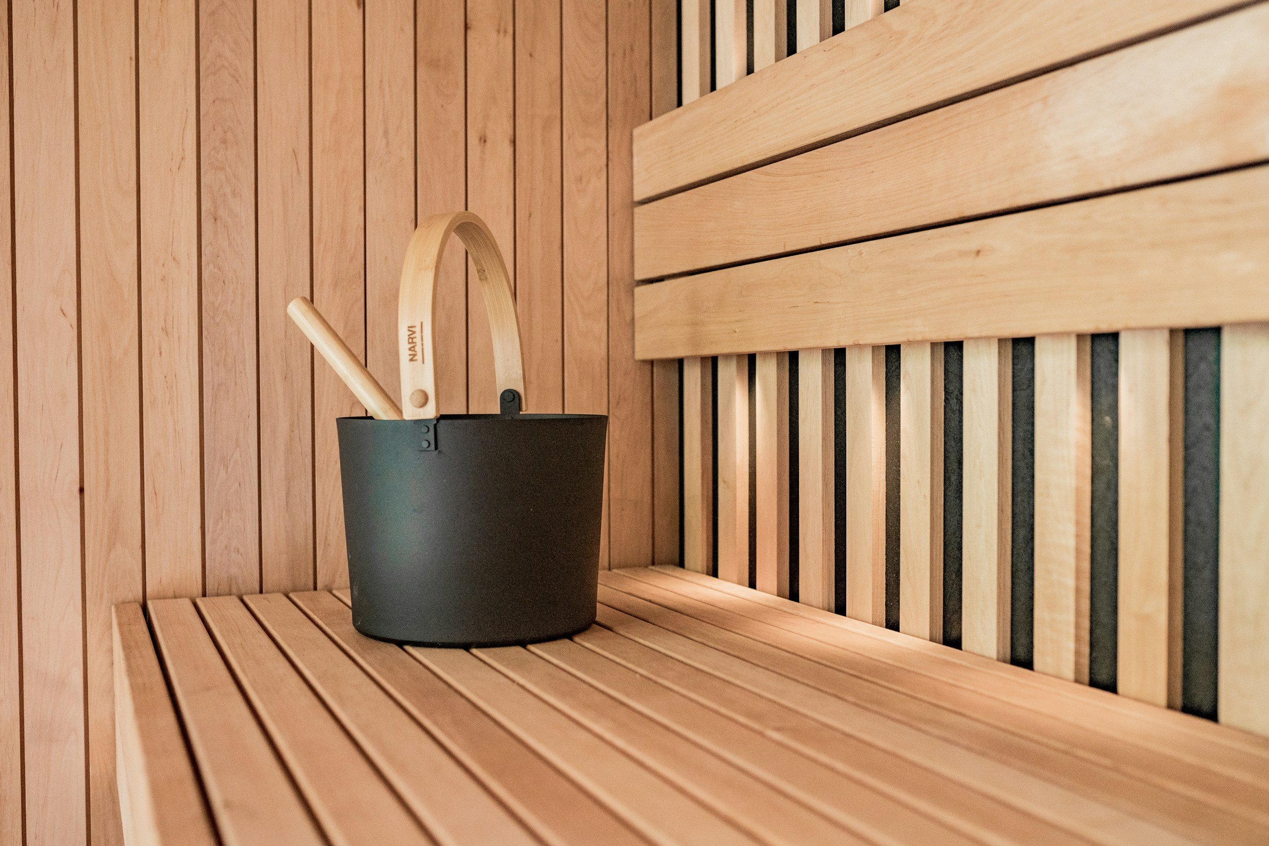 A sauna room with wooden slats on the walls and bench, containing a small black sauna bucket with a wooden ladle resting inside.