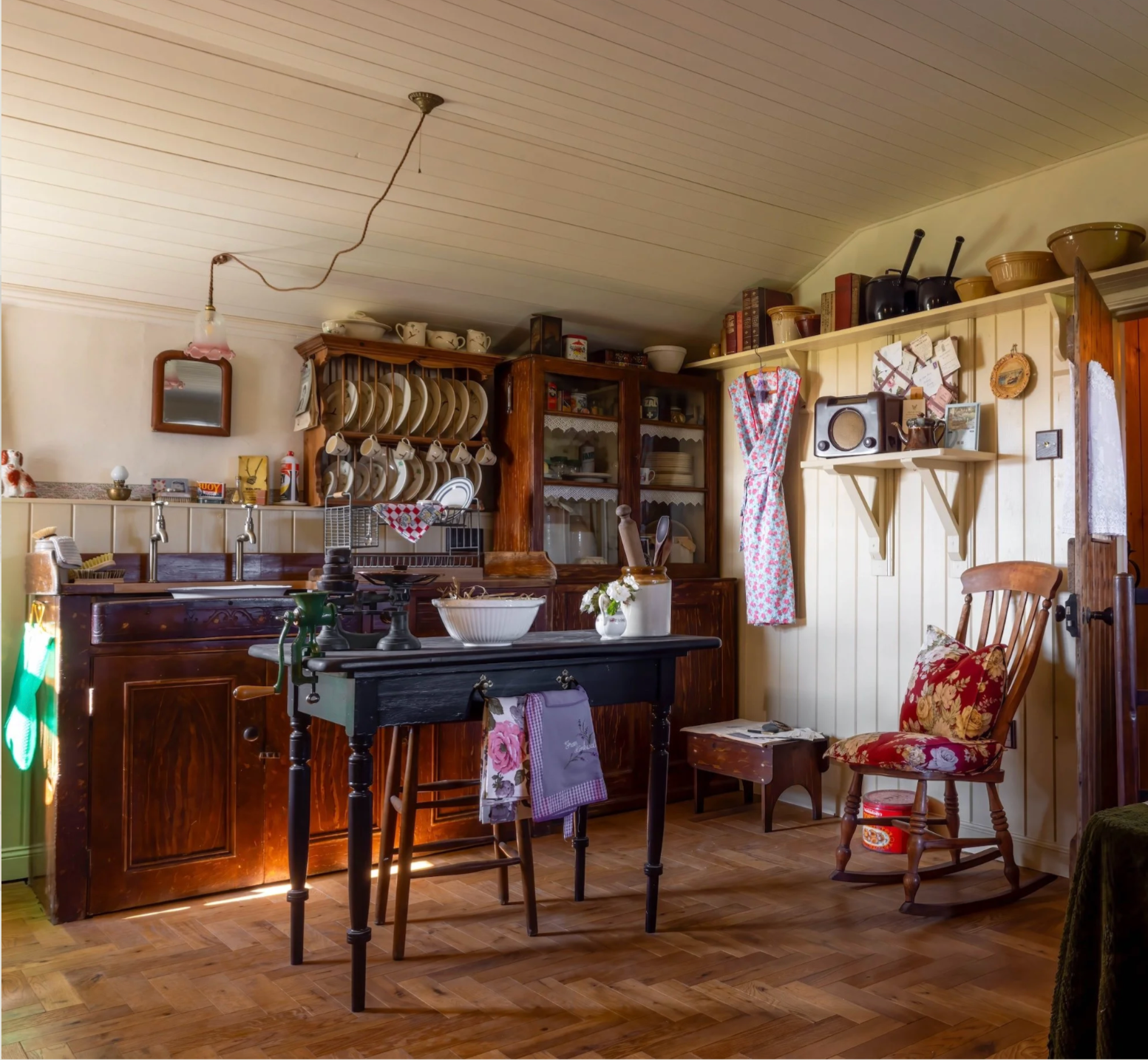 Cozy rustic kitchen with wooden cabinets, shelves, and a wooden chair with floral cushion. Dishes and bowls are on the shelves, and a small table with kitchen utensils is in the center. A curtain covers a window, and various kitchen items are visible