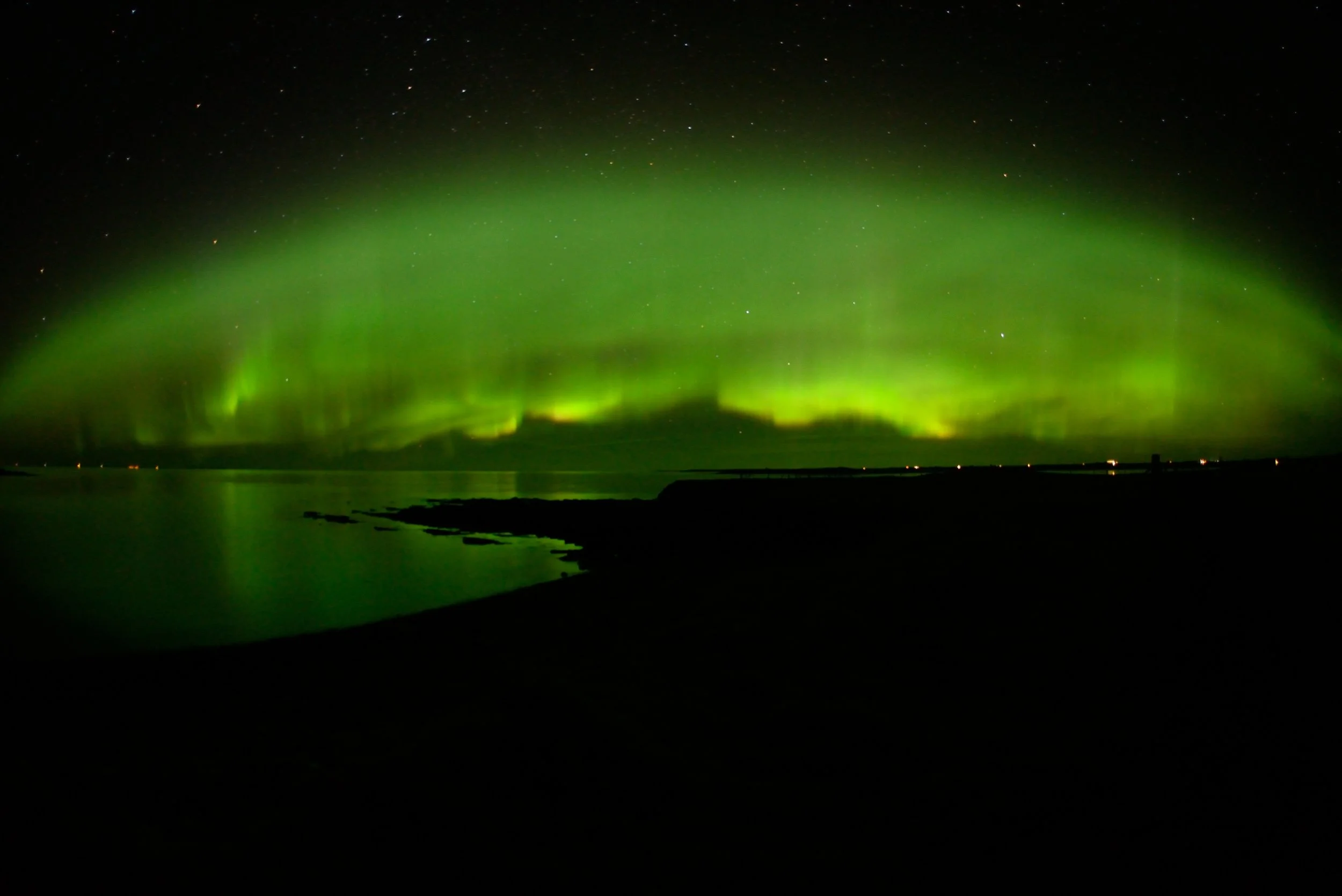 Green aurora borealis illuminating the night sky over a dark shoreline with calm water and distant lights on the horizon.