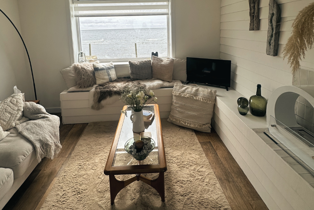 Cozy living room with a window seat, a beige sofa with throw blankets, a wooden coffee table with a vase of flowers, a small television, and decorative vases on a white built-in shelf, with natural light streaming through the window.