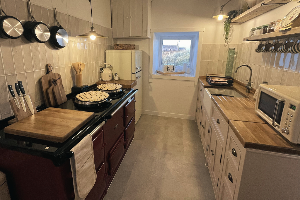 A cozy kitchen with beige tiled walls, wooden countertops, and white cabinets. Contains a vintage red stove with pots on top, a white refrigerator, a microwave, and various kitchen utensils. A window shows an outdoor view.