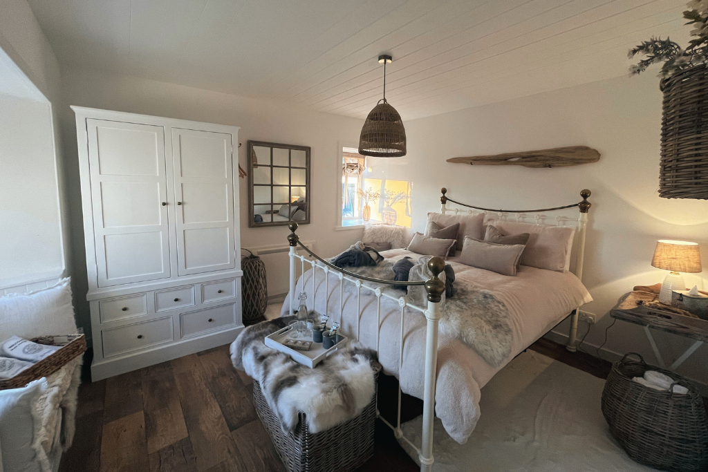 Cozy bedroom with white iron bed, multiple pillows, and fur throw blanket. Nightstand with lamp, woven baskets, wooden shelf on wall, and window with natural light.