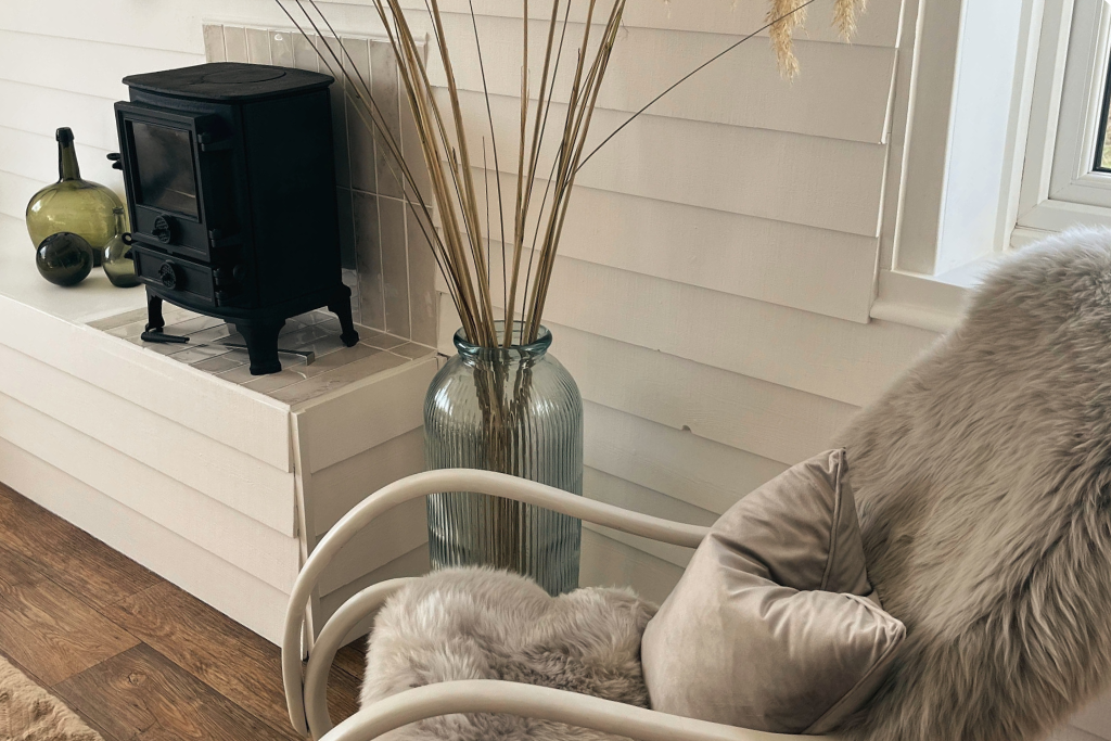 A cozy corner with a white armchair, gray fluffy pillow, and fur blanket, next to a large glass vase filled with dried pampas grass, in front of a window and a white wall with horizontal paneling. A small black wood stove and green glass vases are on