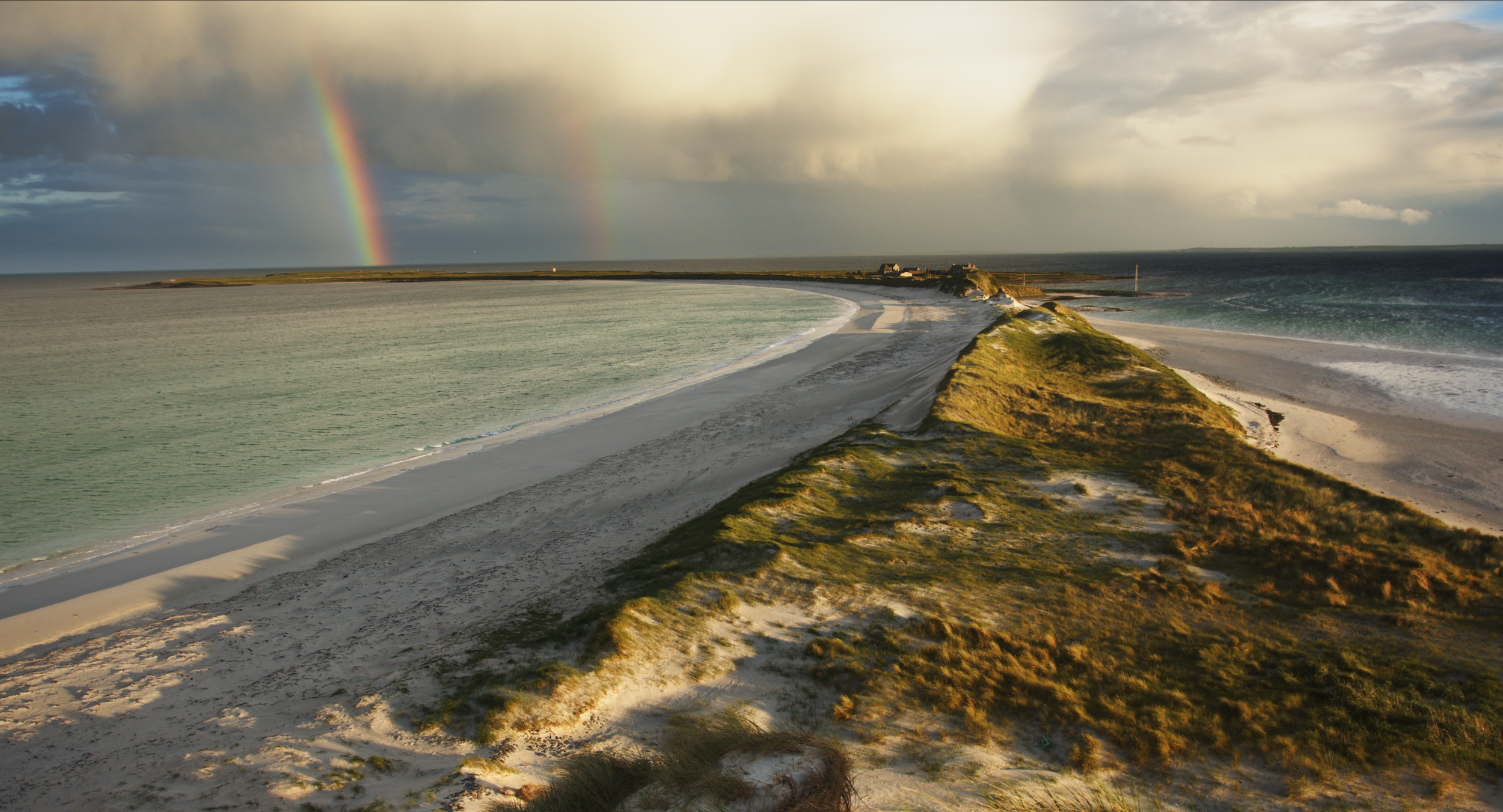 Sanday beach scene during sunset with a sandbar running into the ocean, green grassy dunes, and a rainbow in the sky with dark clouds.