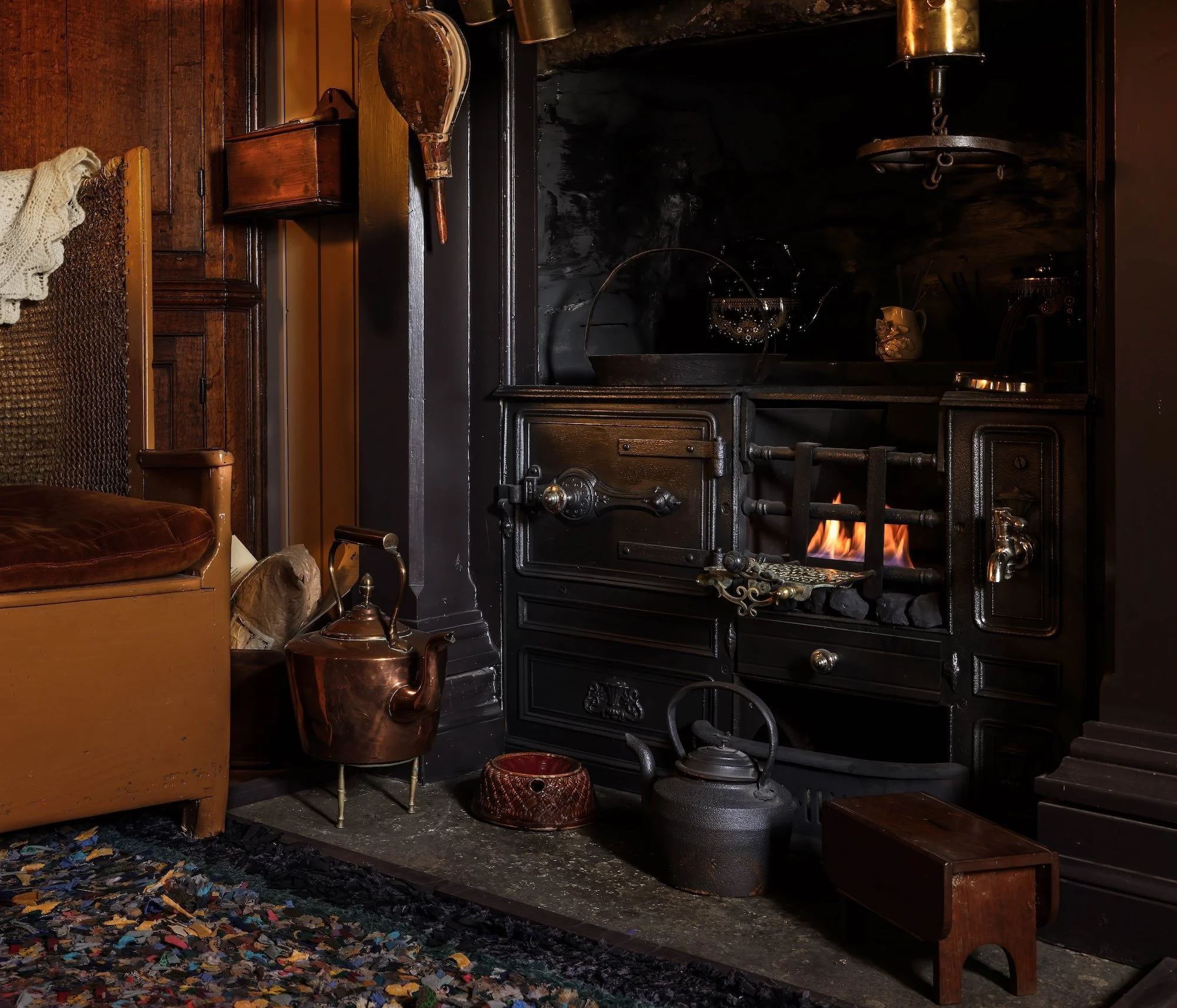 A cozy vintage room in Boloquoy featuring a black cast iron stove with a small fire burning inside, surrounded by various antique items including a copper kettle, a wooden box, and a woven basket, with a colorful rug on the floor.