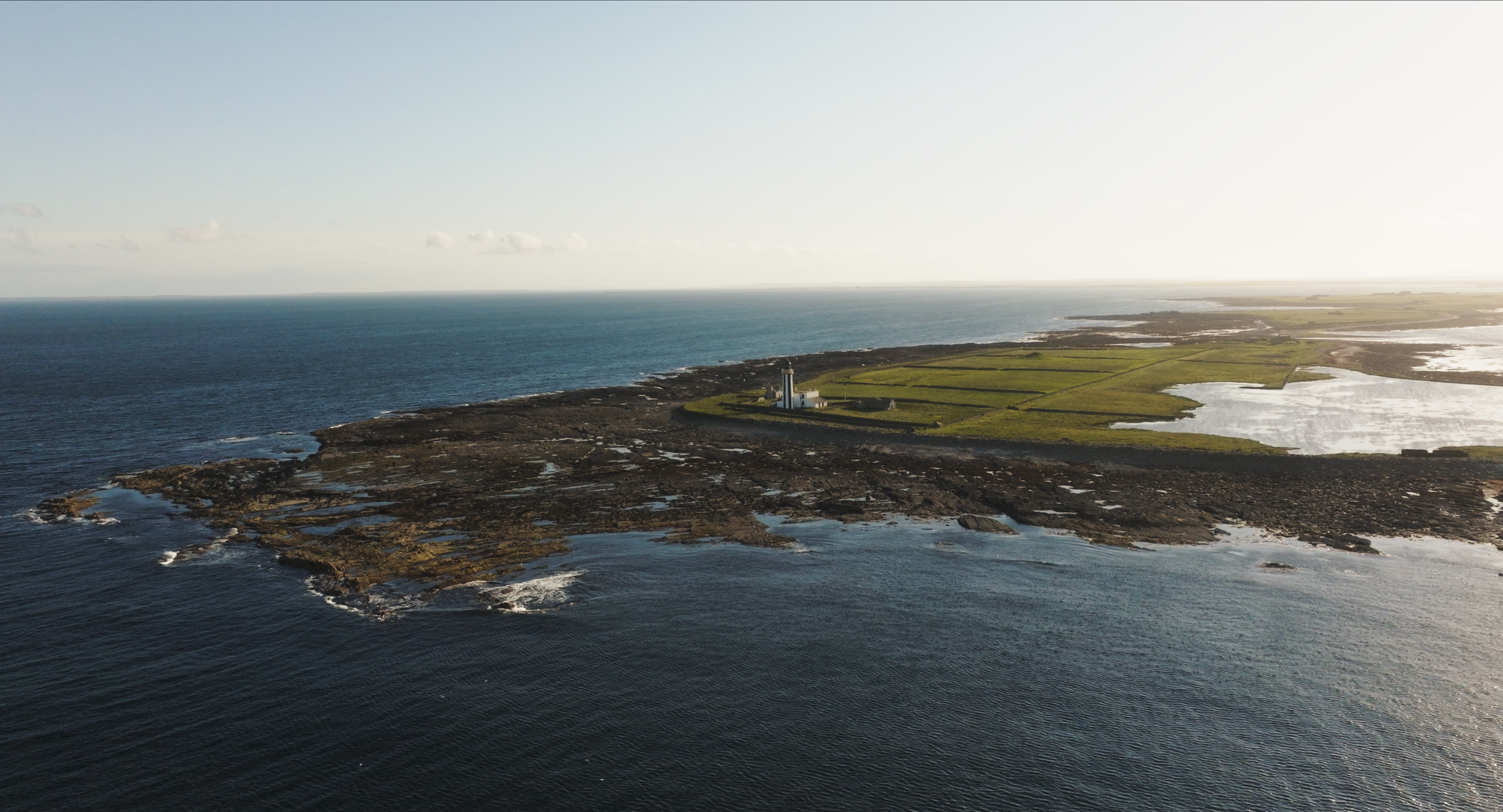 Aerial view of Sanday, a rocky coastline with a lighthouse, green fields, and ponds, with the ocean on the left and the sun setting on the right.