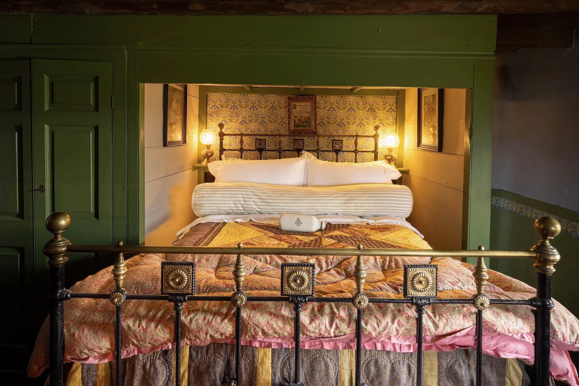 A cozy bedroom with a vintage-style iron bed frame, decorated with a floral quilt and multiple pillows. The headboard is positioned in a small alcove with wallpapered walls, illuminated by two bedside lamps. The room has green wooden walls and framed artwork.