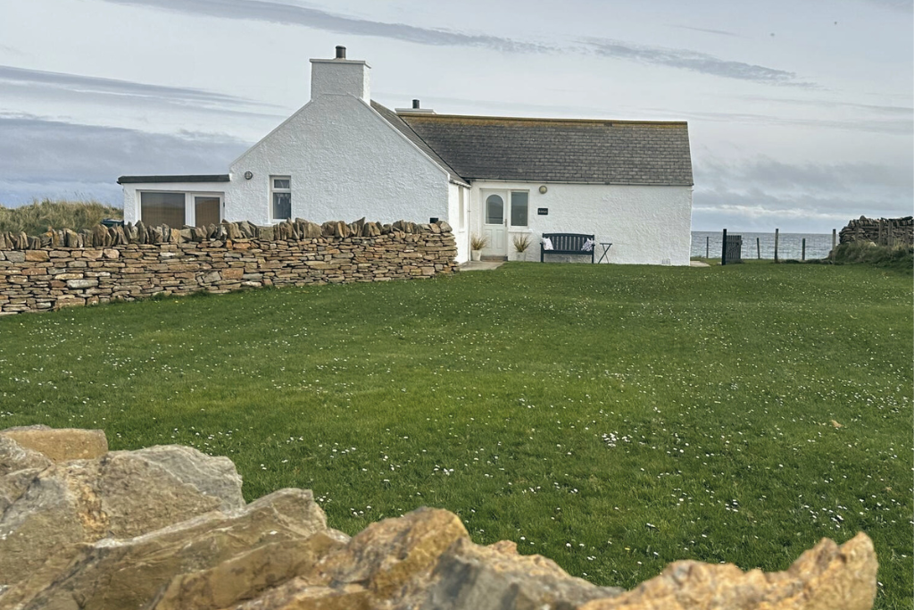 Kilnbarn Cottage, a white house with a sloped roof and chimney, surrounded by a stone wall and green lawn, near the ocean under a cloudy sky.