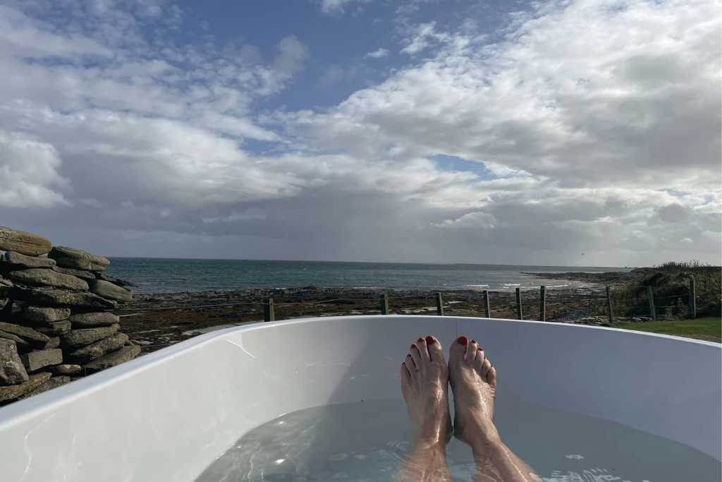 Person relaxing in a white outdoor bathtub at Kilnbarn cottage, with a view of the ocean and cloudy sky.