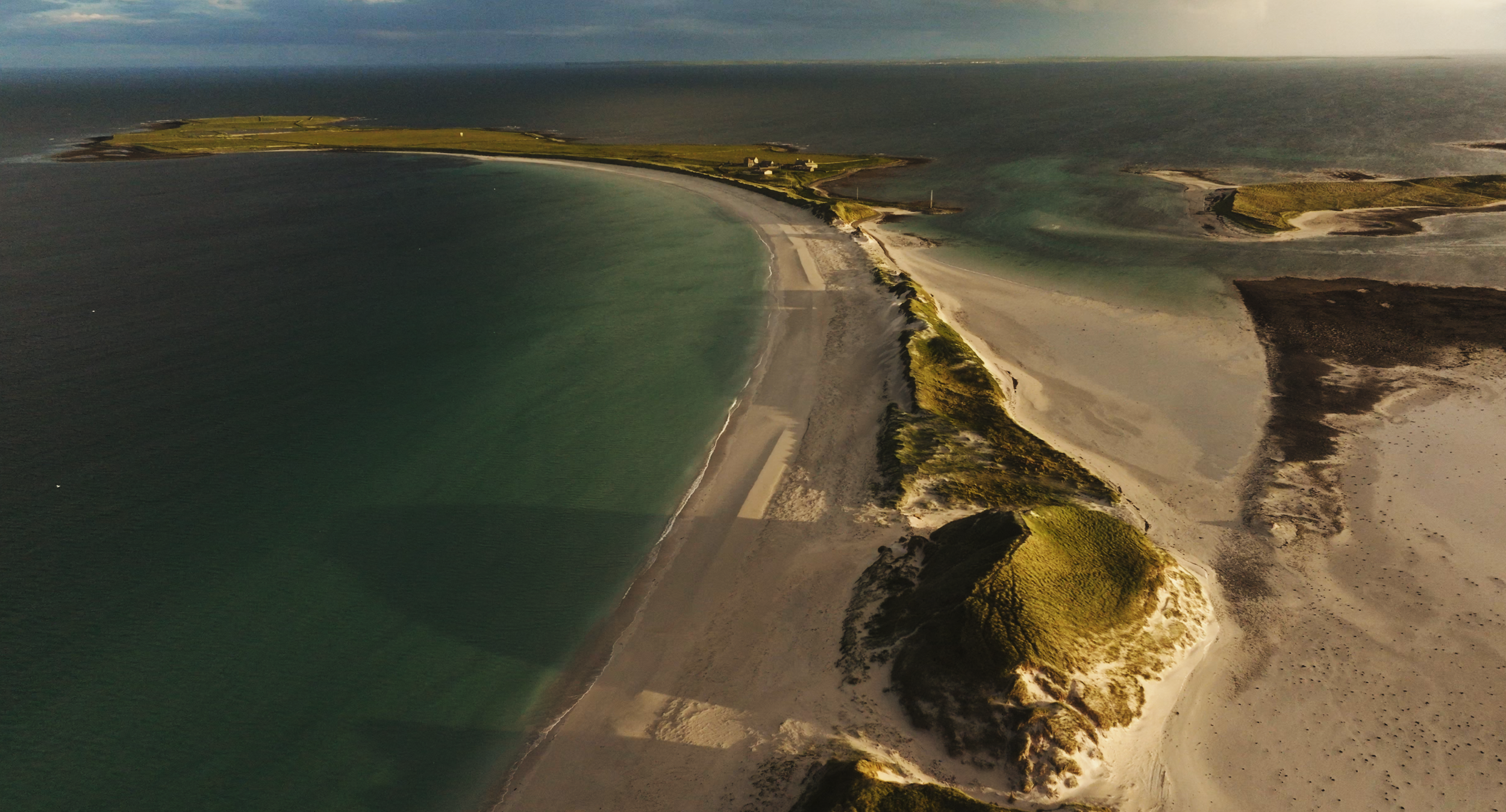 Aerial view of Sanday coastal landscape featuring sandy beaches, green grassy areas, and dark rocky sections, with the ocean on the left and small buildings in the distance.