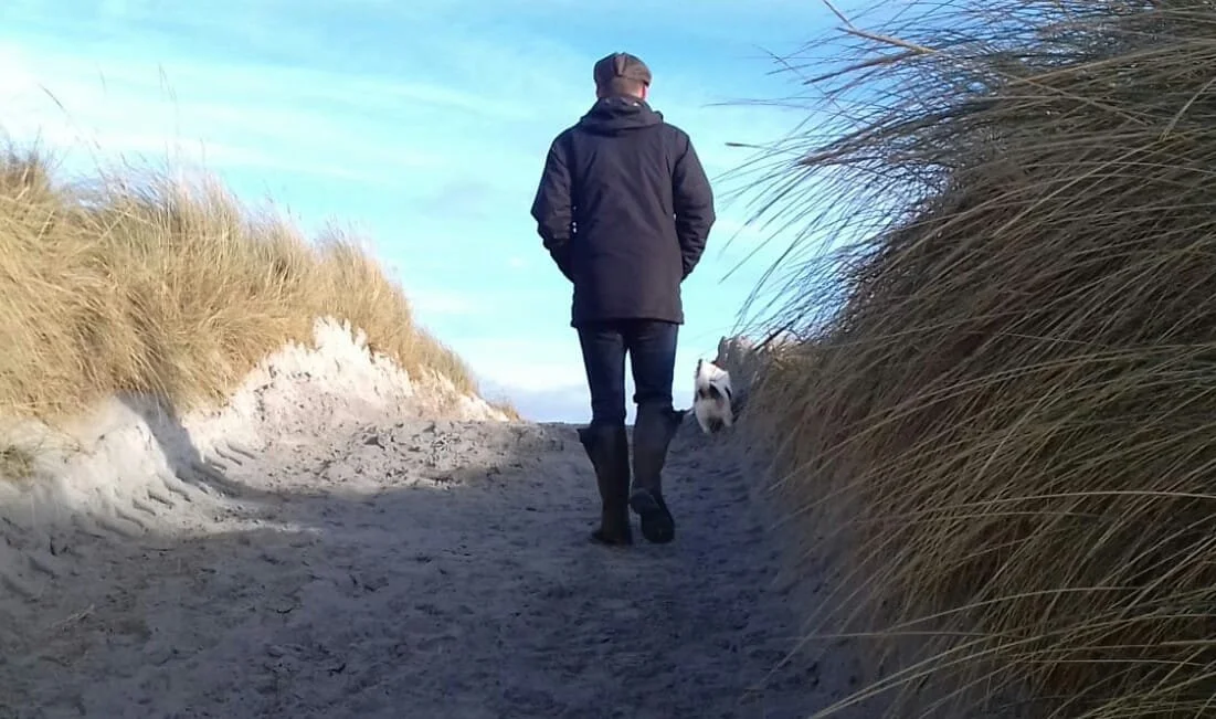 Person walking on a sandy beach path with a small dog, flanked by tall grass on both sides, under a blue sky.