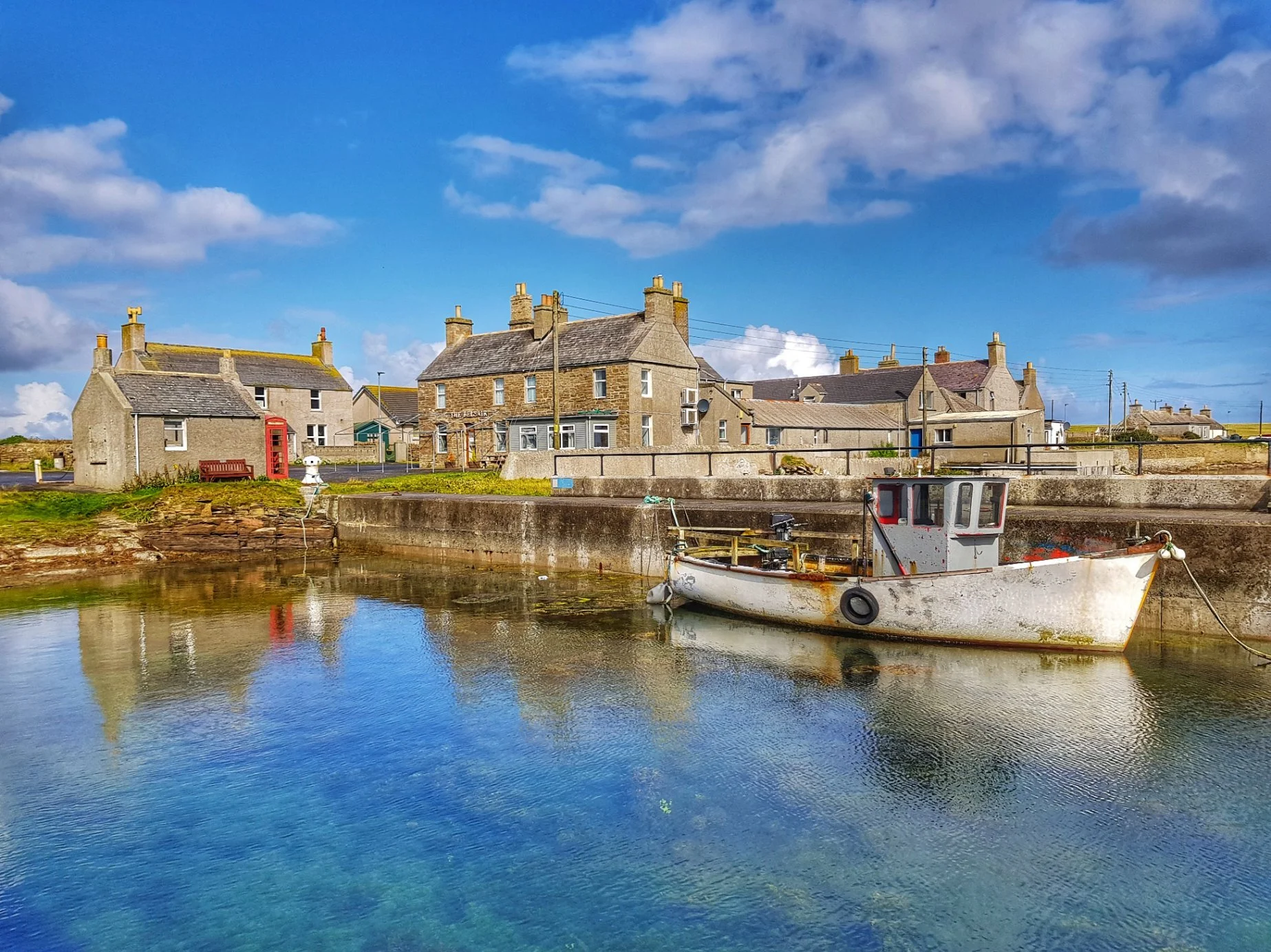 A small fishing boat docked in a harbor at Sanday, with water reflecting the sky and buildings. Behind the boat, there are stone houses with chimneys, a bench, and a red phone booth under a partly cloudy blue sky.